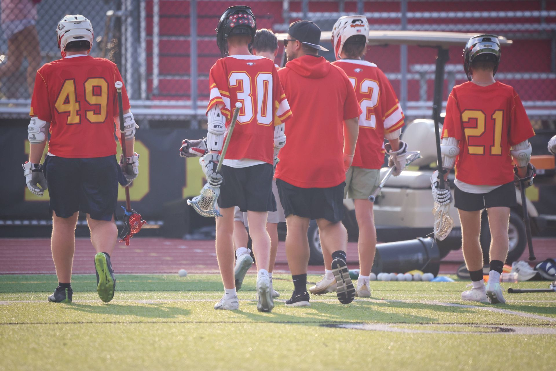 Lacrosse players in red jerseys walk on the field, numbers 49, 30, 32, and 21 visible