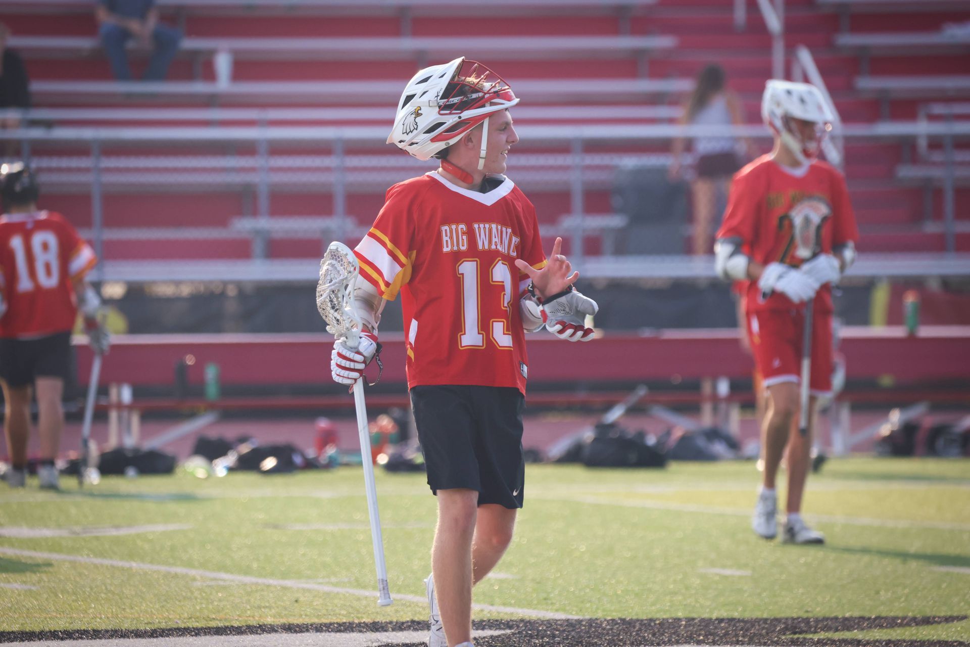 Lacrosse player in red jersey holding a stick on a field with teammates and bleachers in the background