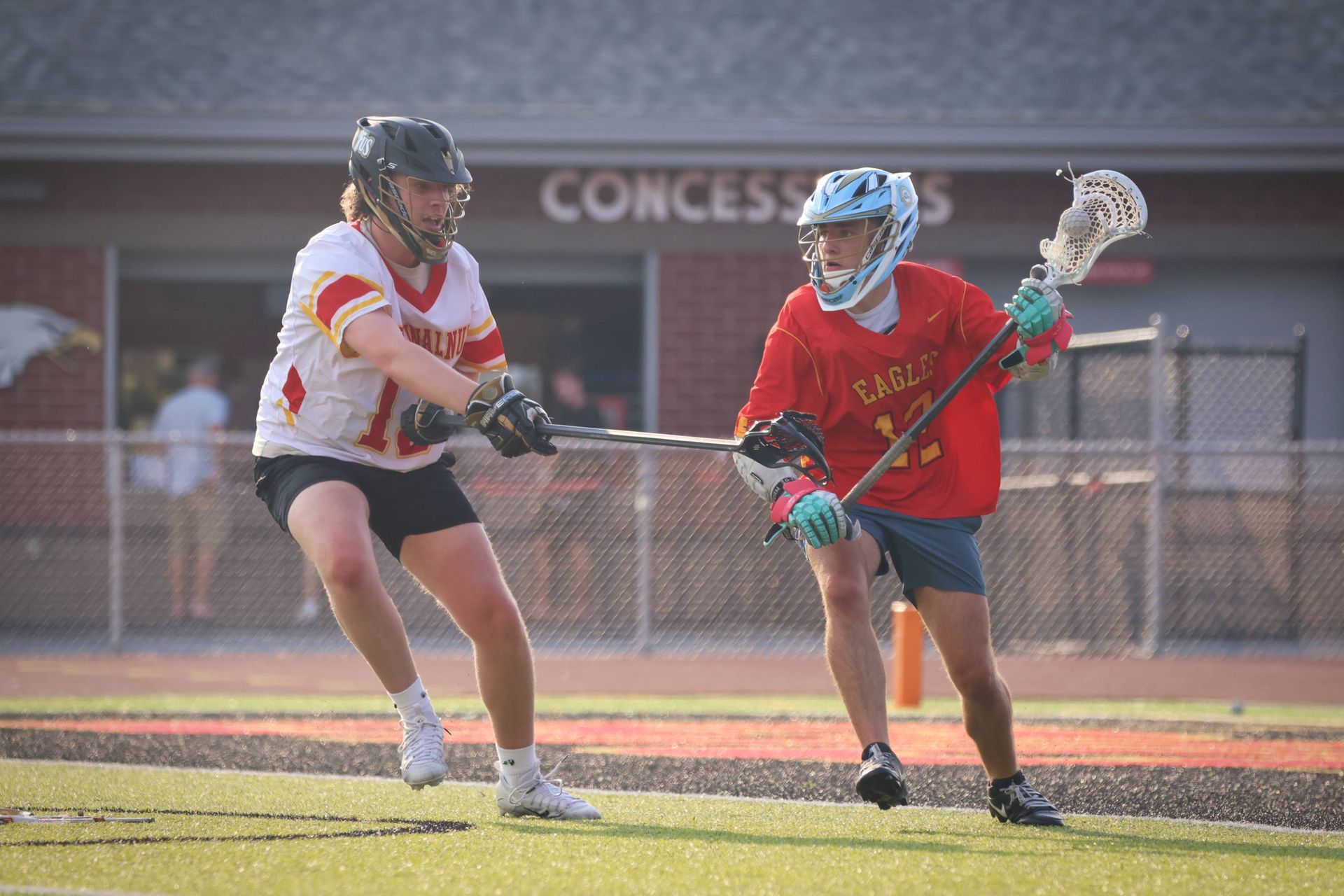 Two lacrosse players in red and white jerseys compete on a field near the goal.