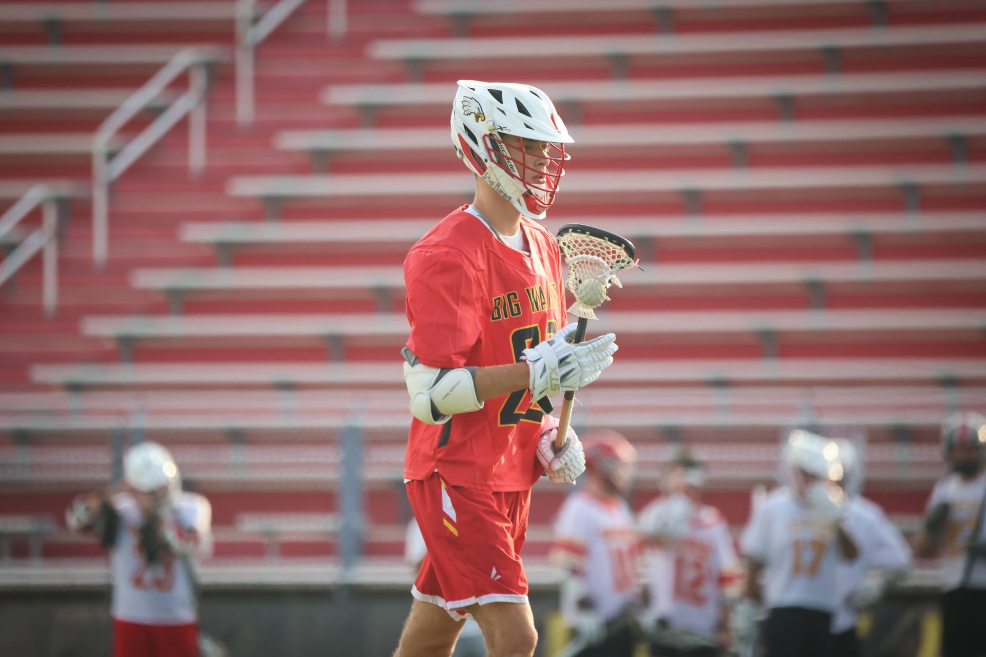 Lacrosse player in red uniform holding a stick on a field with blurred players and stadium bleachers behind him