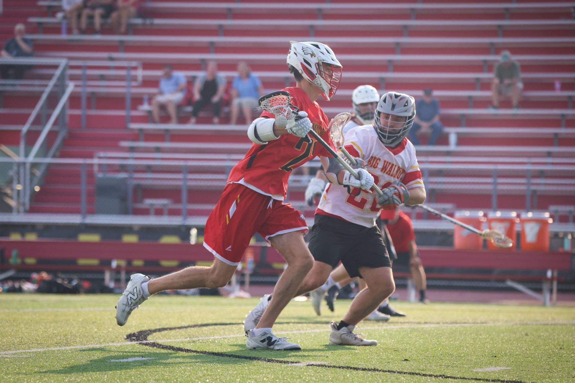 Two lacrosse players in red and white sprint across a field during a game.