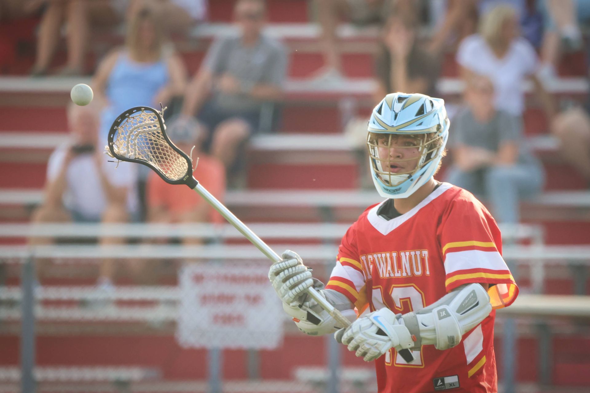 Lacrosse player in orange jersey and blue helmet holding a stick on the field with bleachers behind him