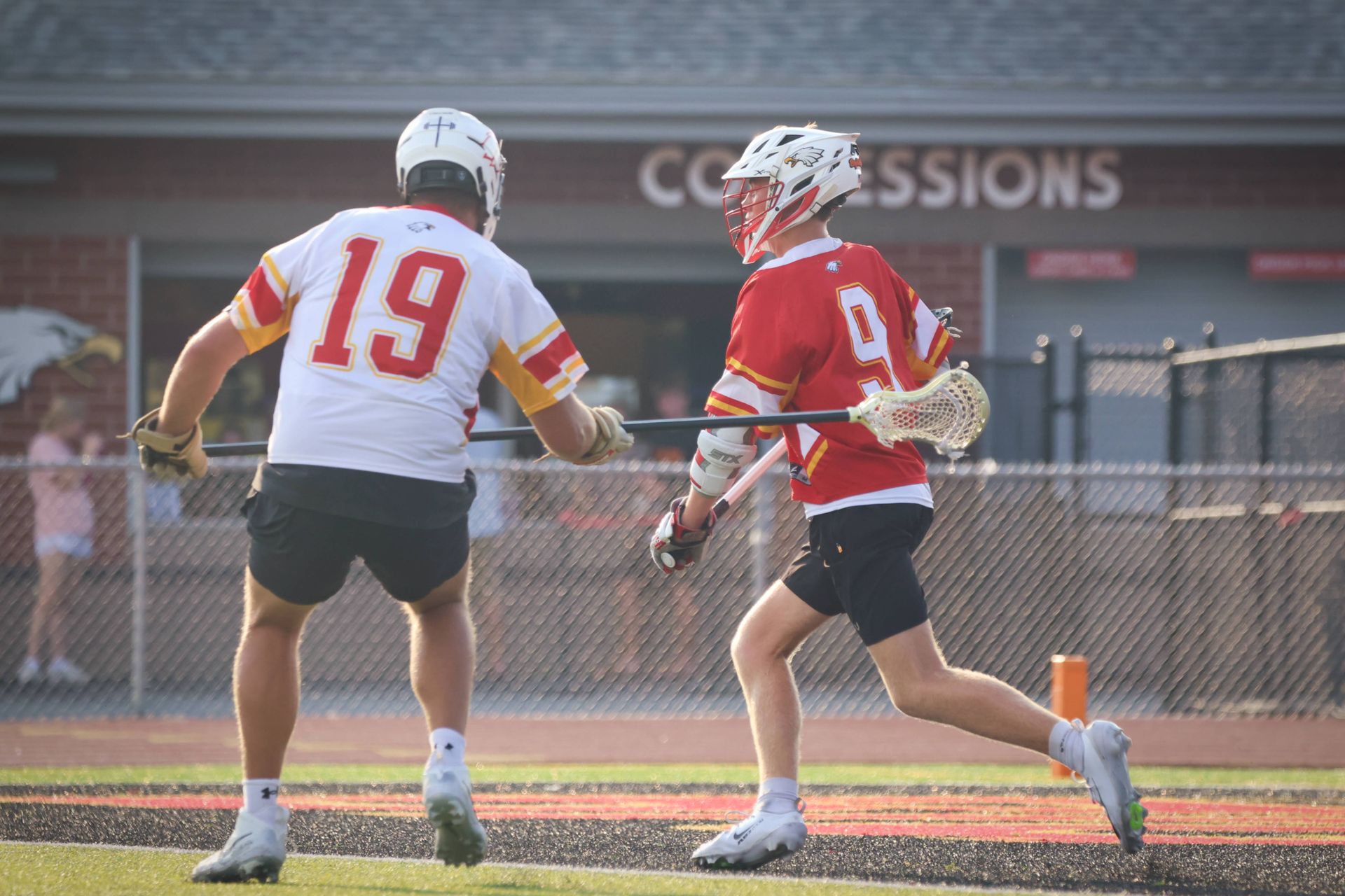Two lacrosse players in red and white uniforms on a field, one holding a stick near a goal