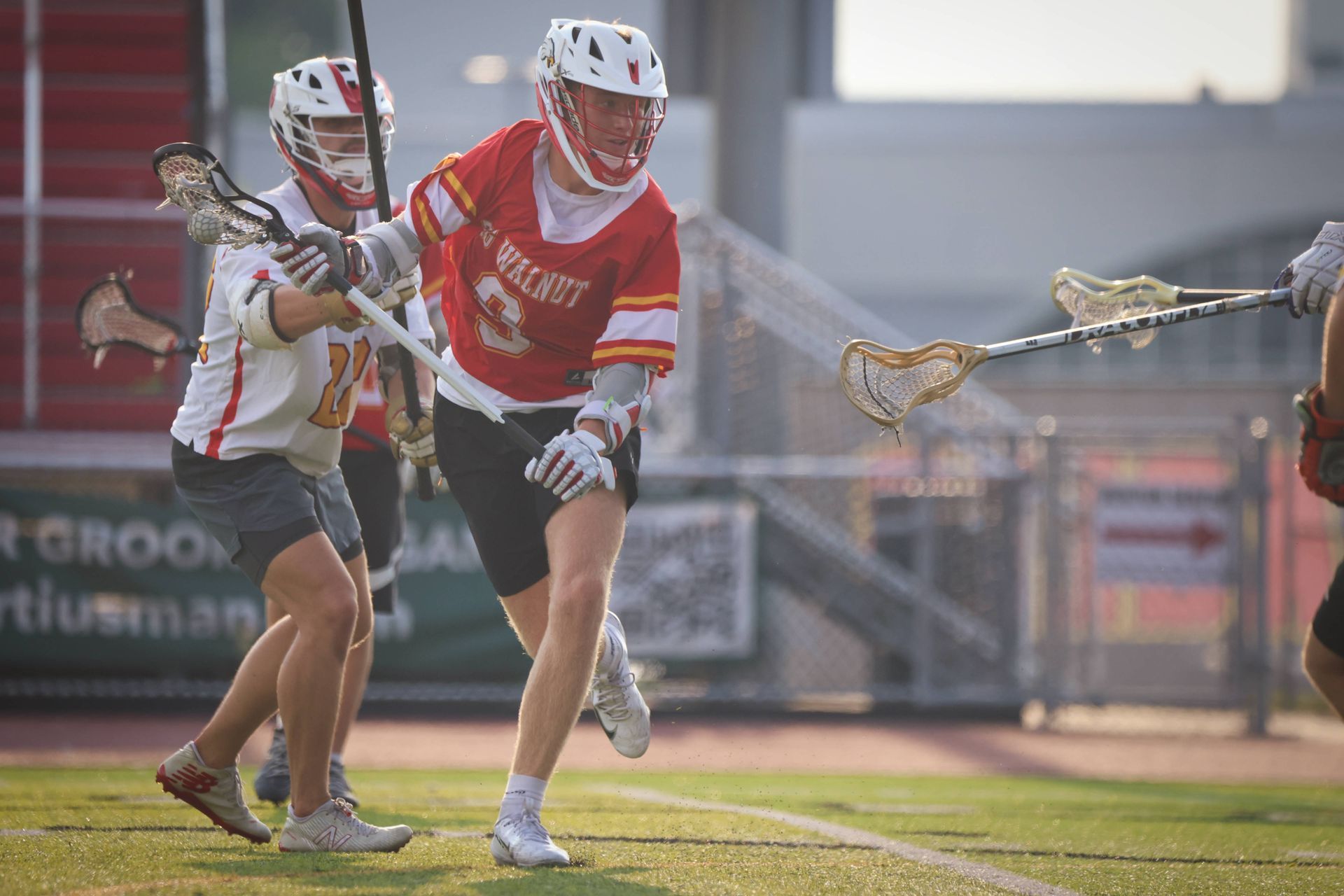 Two lacrosse players in red and white uniforms sprint on a field during a game