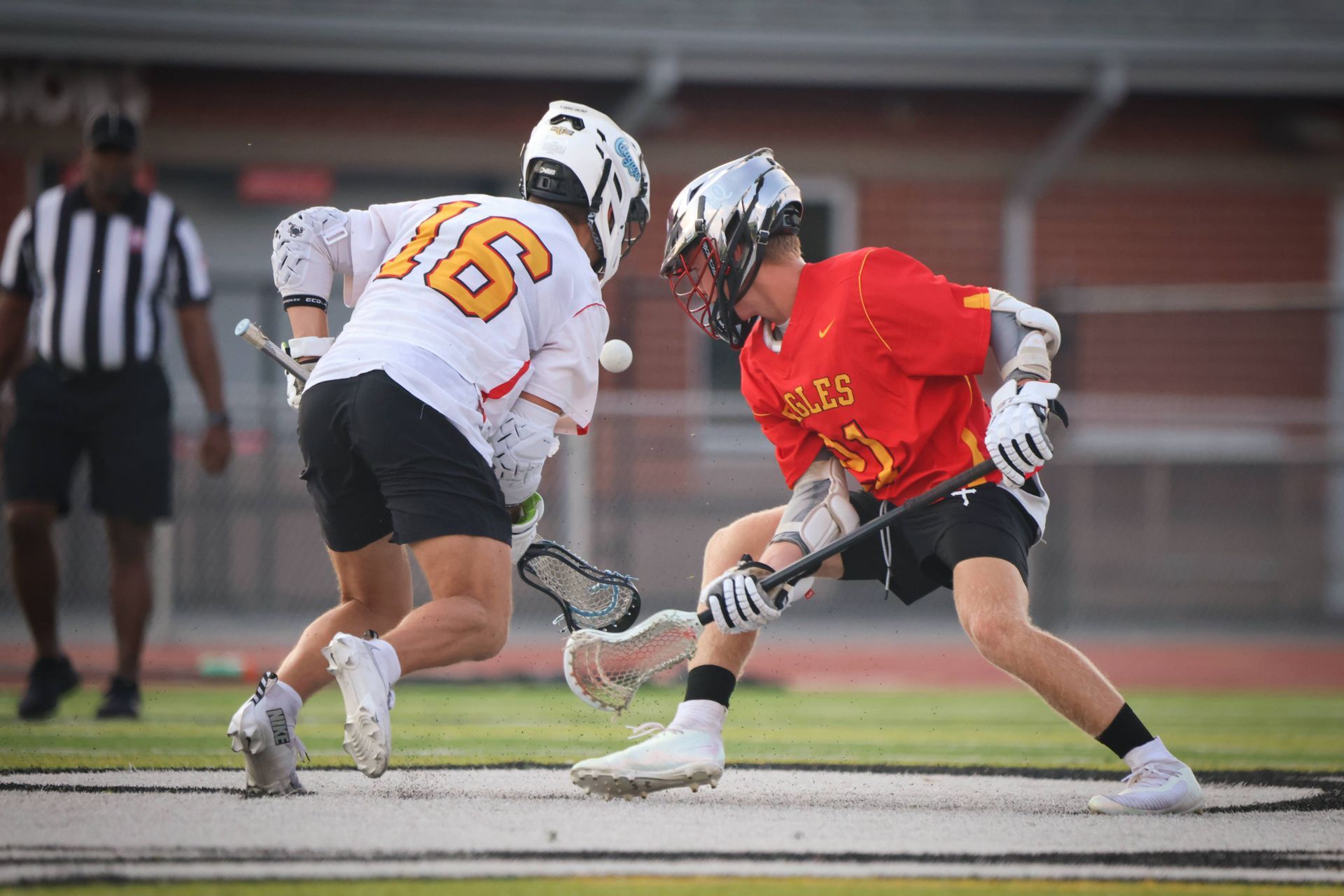 Two lacrosse players in red and white jerseys battle for the ball on a field