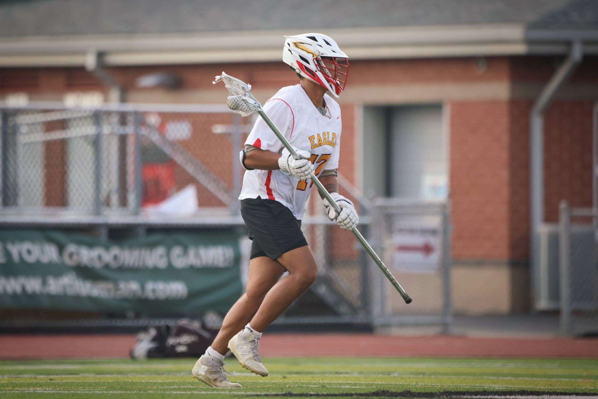 Lacrosse player in white jersey running on a field, carrying a long stick near a brick building