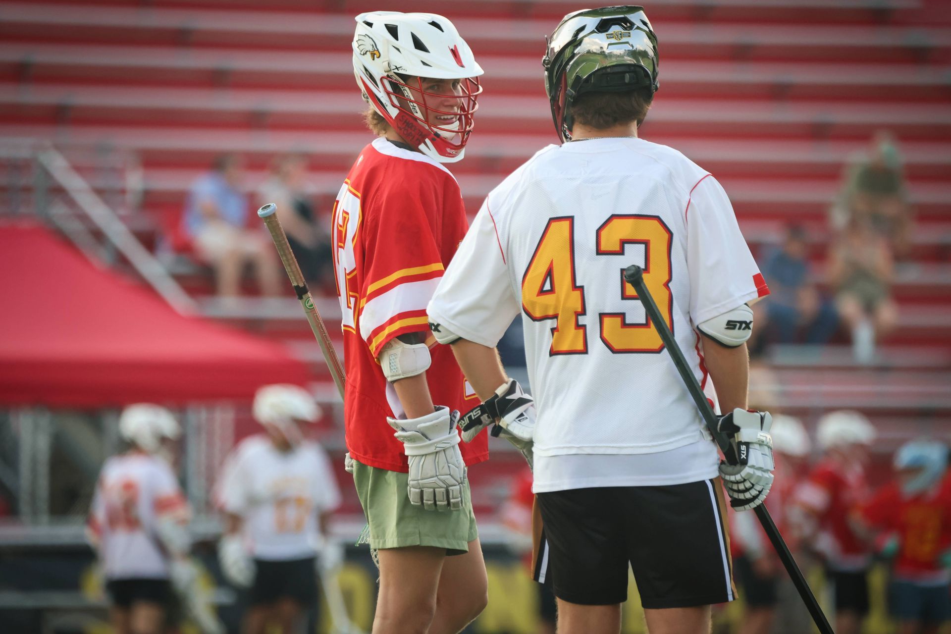 Two lacrosse players in red and white jerseys stand on a field with bleachers in the background.