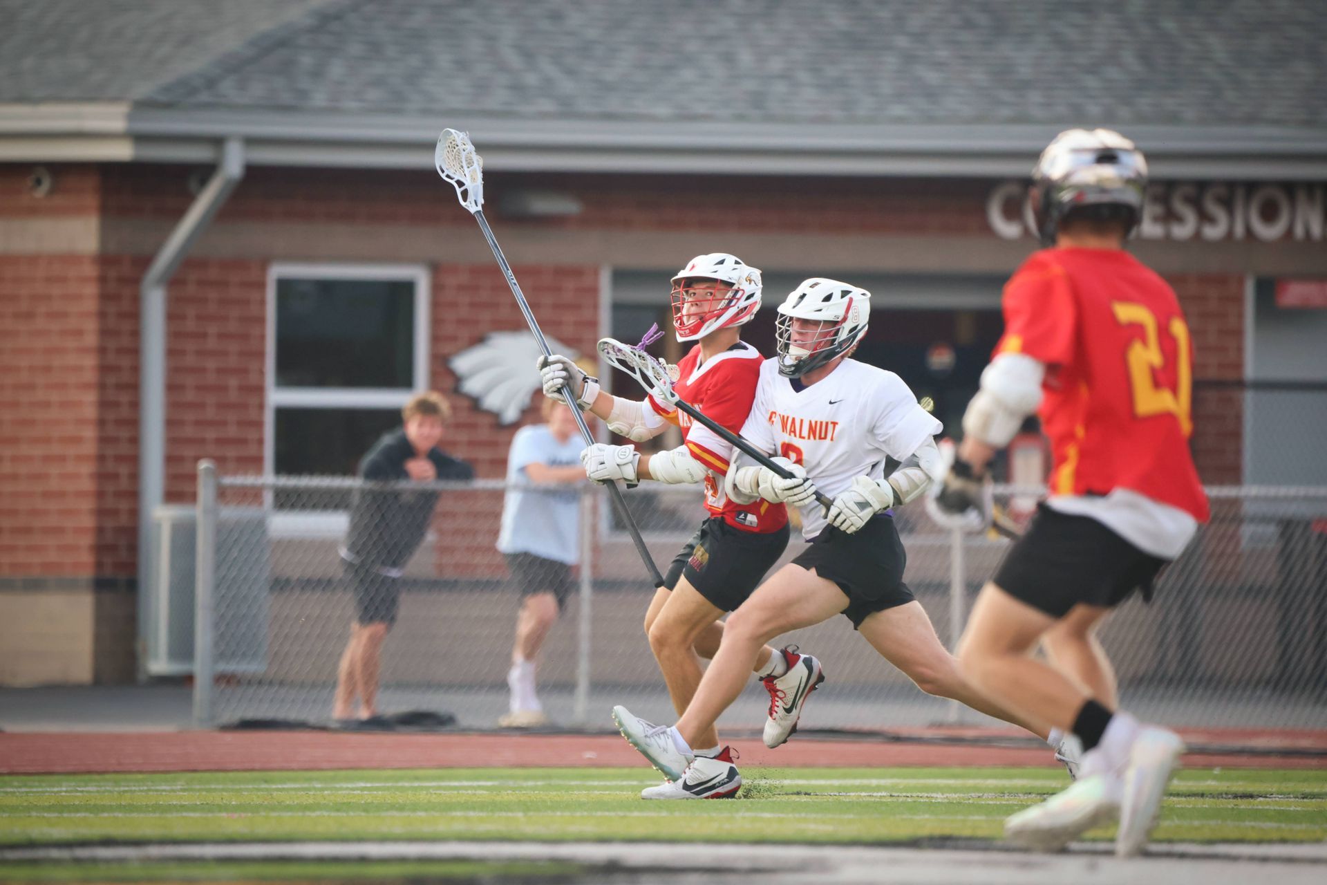 Lacrosse players in red and white sprint across a field during a game, with spectators in the background.