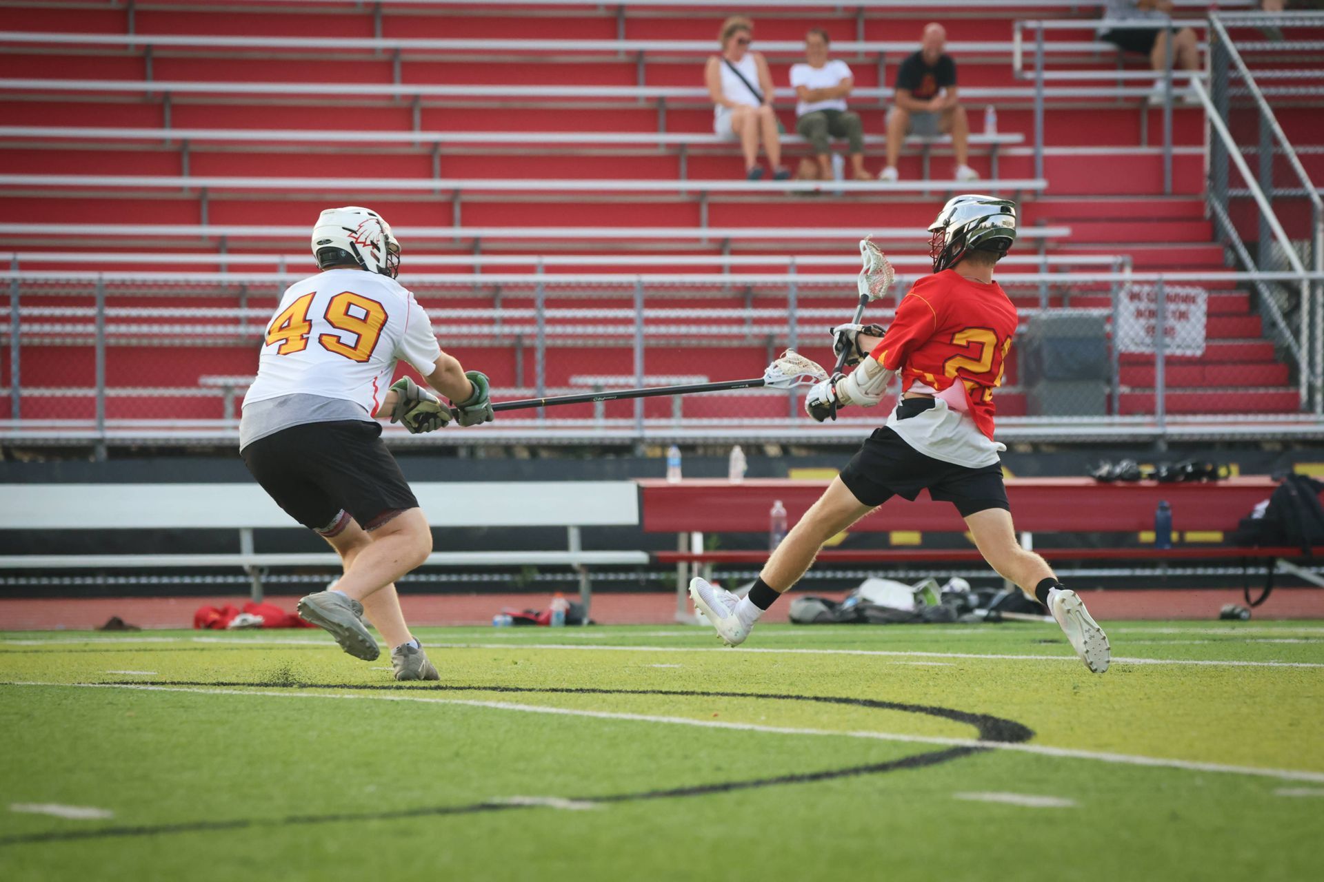 Two lacrosse players sprint on a field, one in white and black, one in red and black, with bleachers behind them