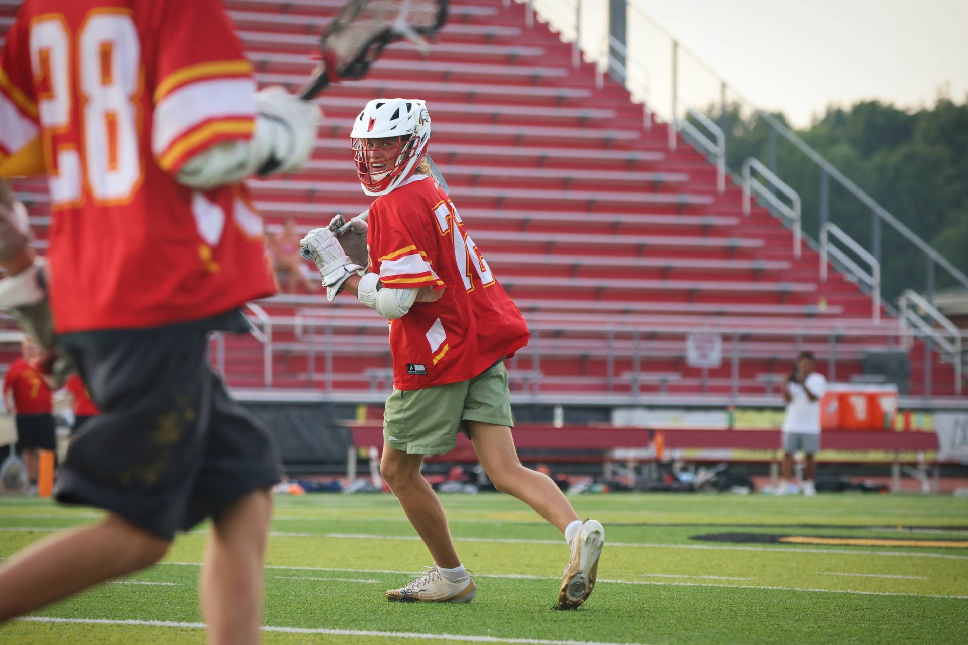 Two lacrosse players in red jerseys on a field, one running with the ball near bleachers.
