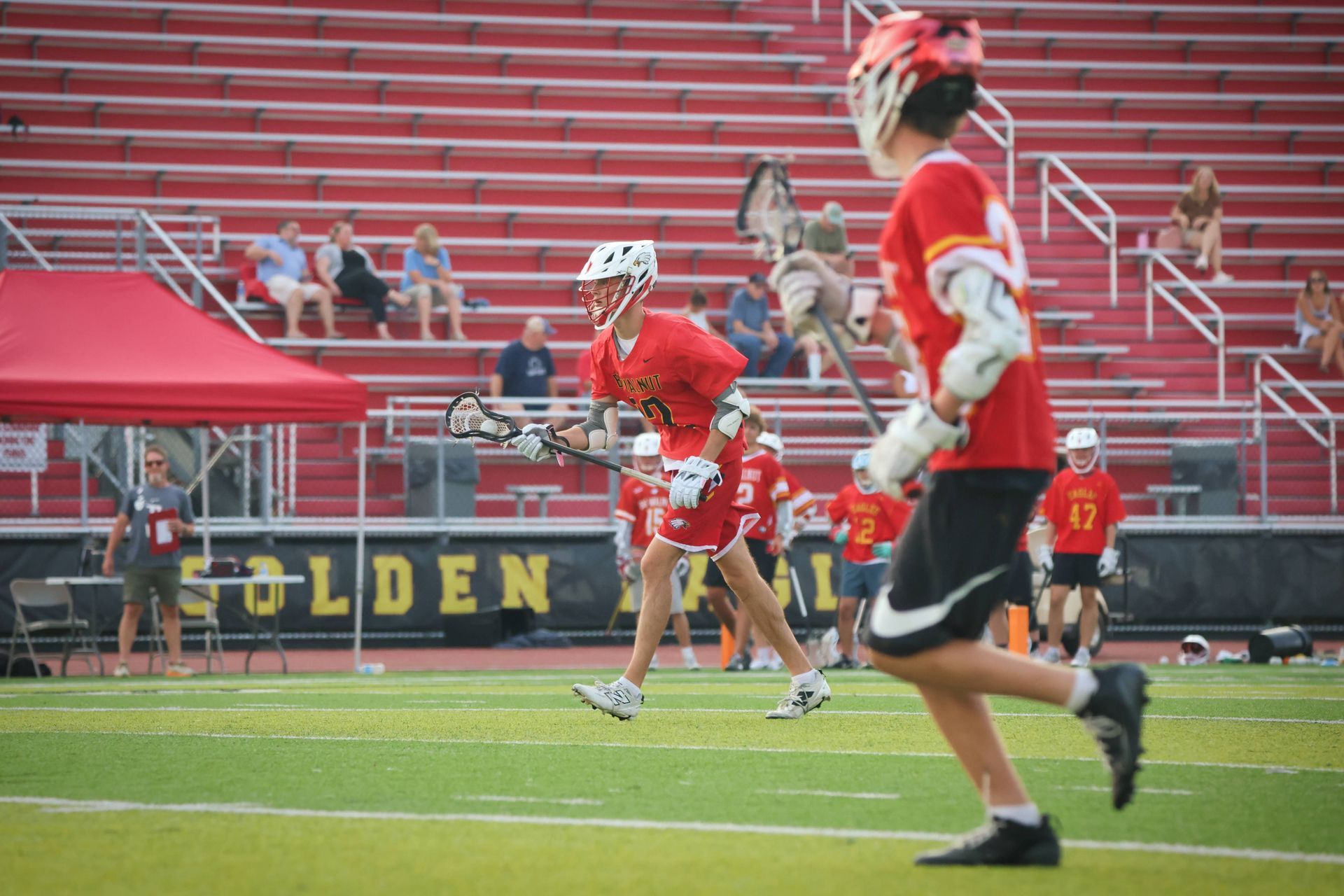 Lacrosse players in red uniforms on a field, with bleachers and spectators in the background.