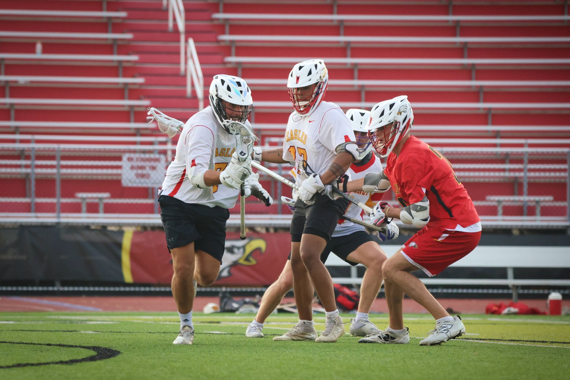 Lacrosse players in white and red uniforms competing on a field with red stadium seats in the background