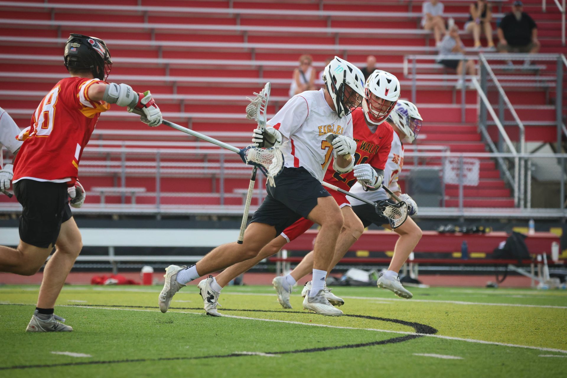 Lacrosse players in red and white sprint across the field during a game, with bleachers in the background.