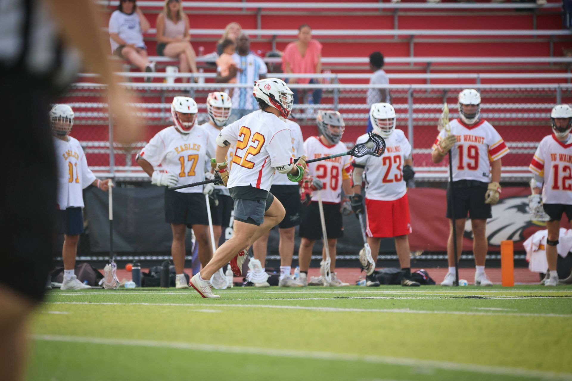 Lacrosse players in white and red uniforms line up on a field near red bleachers.