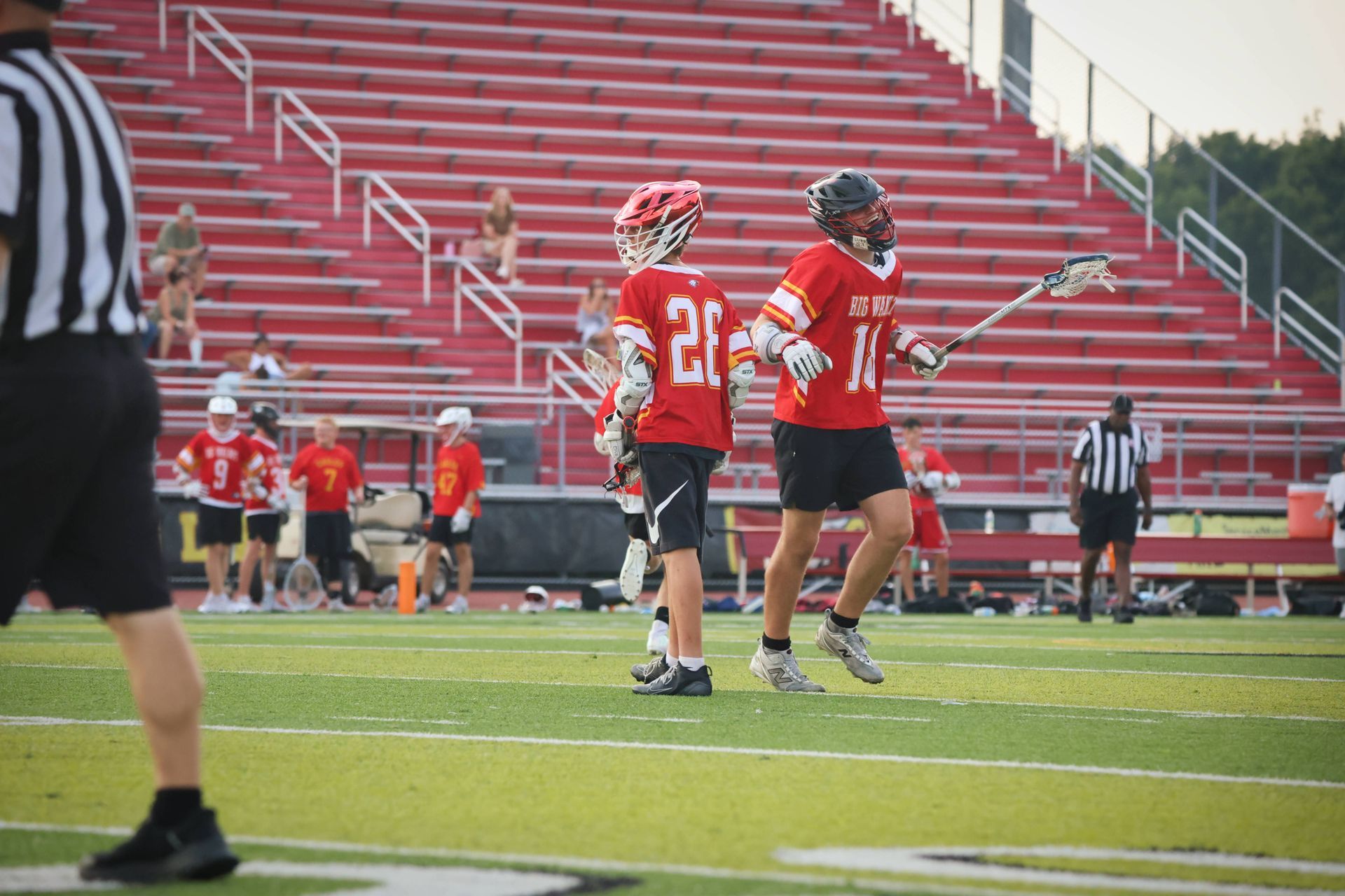 Lacrosse players in red uniforms on a field with empty red bleachers in the background