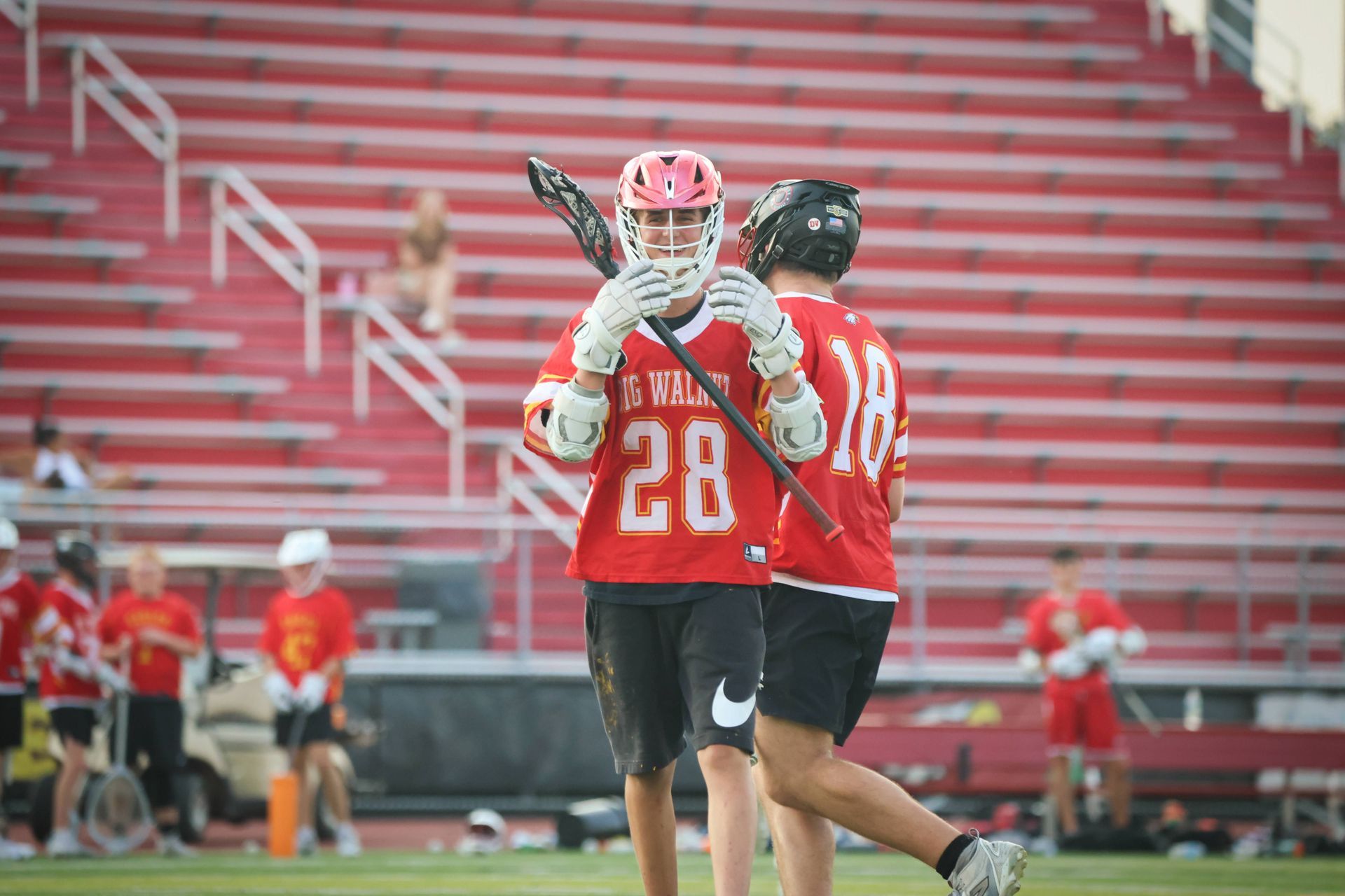 Two lacrosse players in red jerseys celebrate on a field with empty red stadium seats behind them.