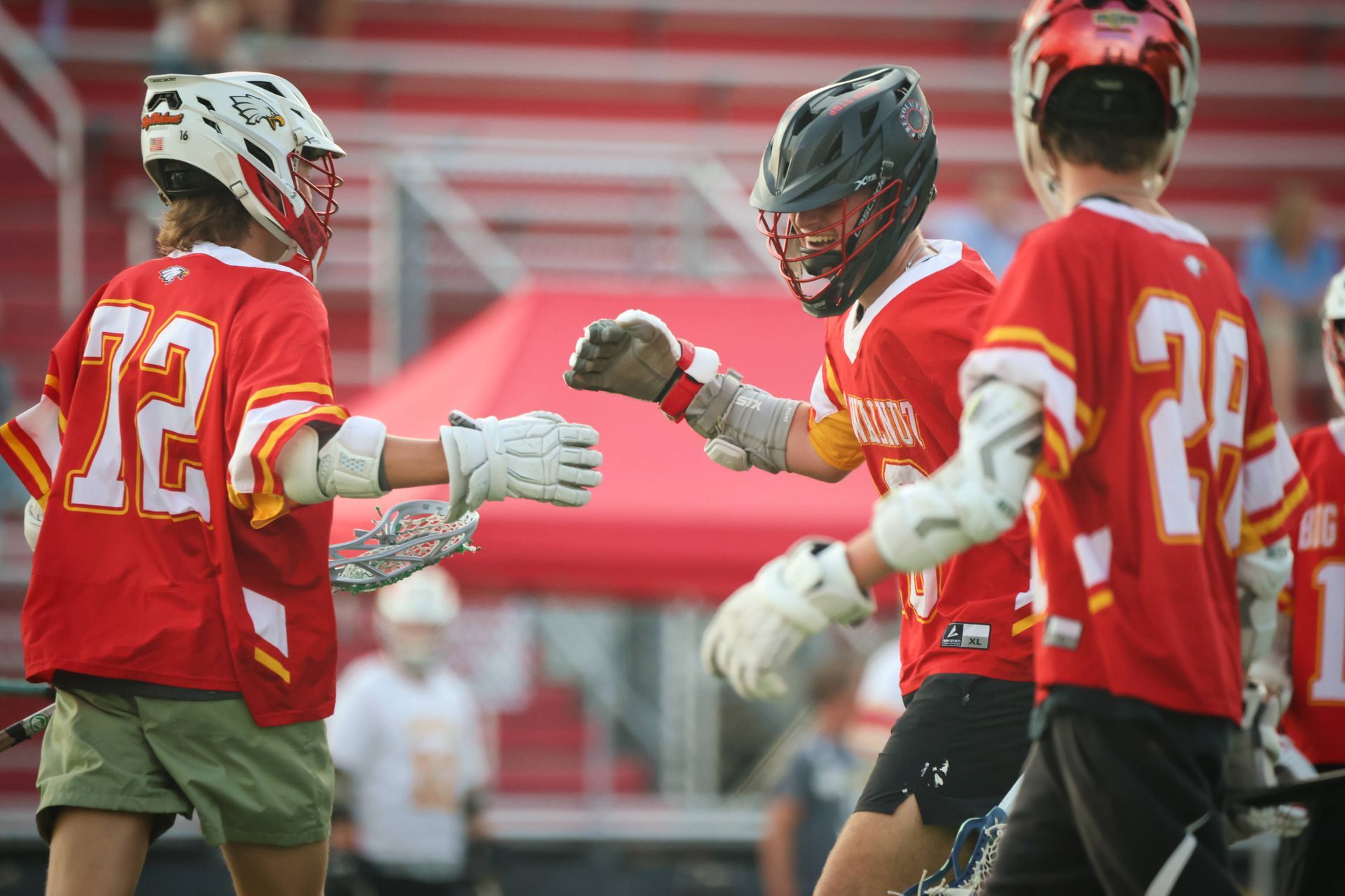 Three lacrosse players in red uniforms huddle on the field, raising gloved fists for a team cheer.