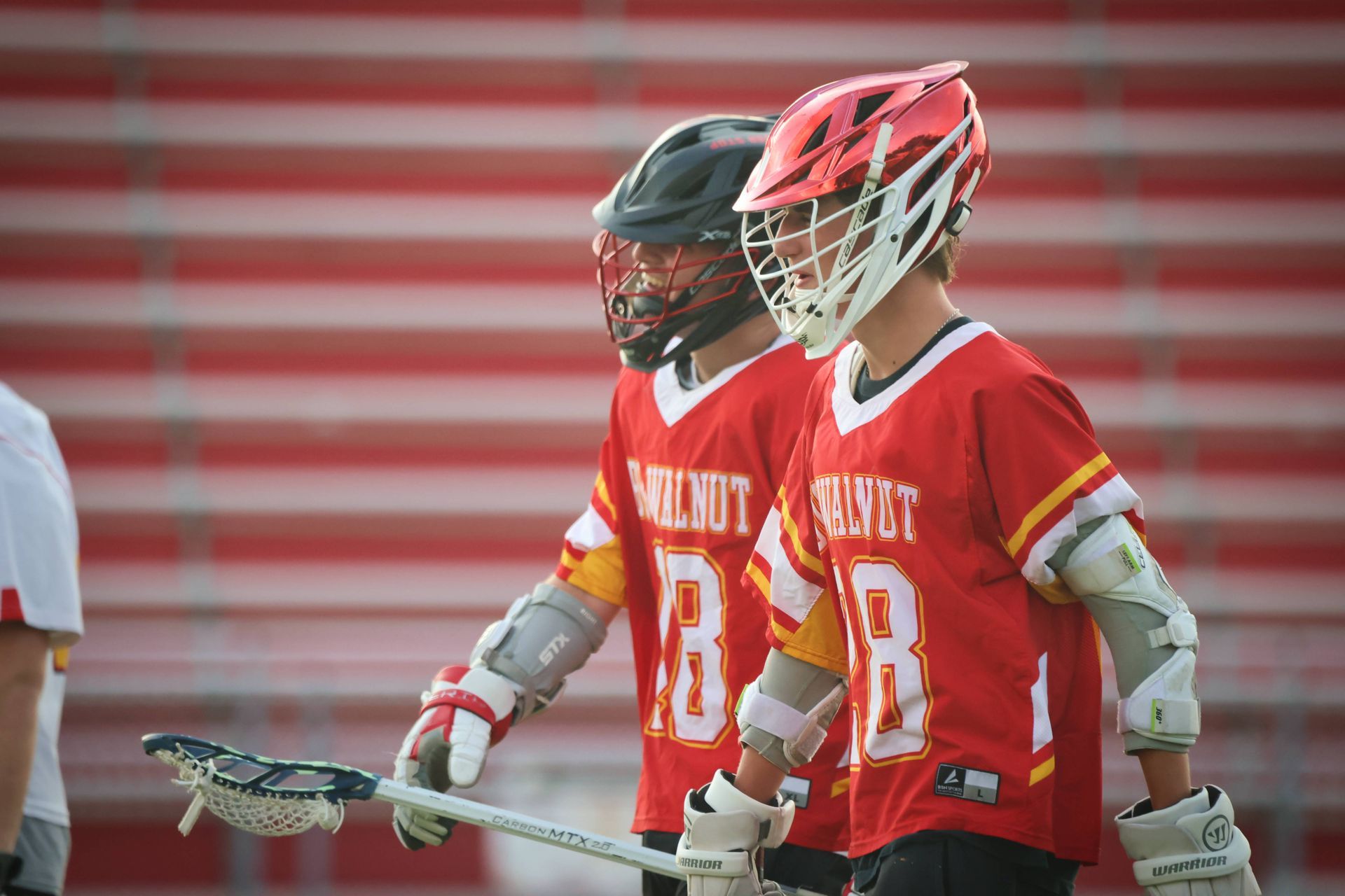 Two lacrosse players in red jerseys and helmets standing on a field with sticks, one holding a gold stick