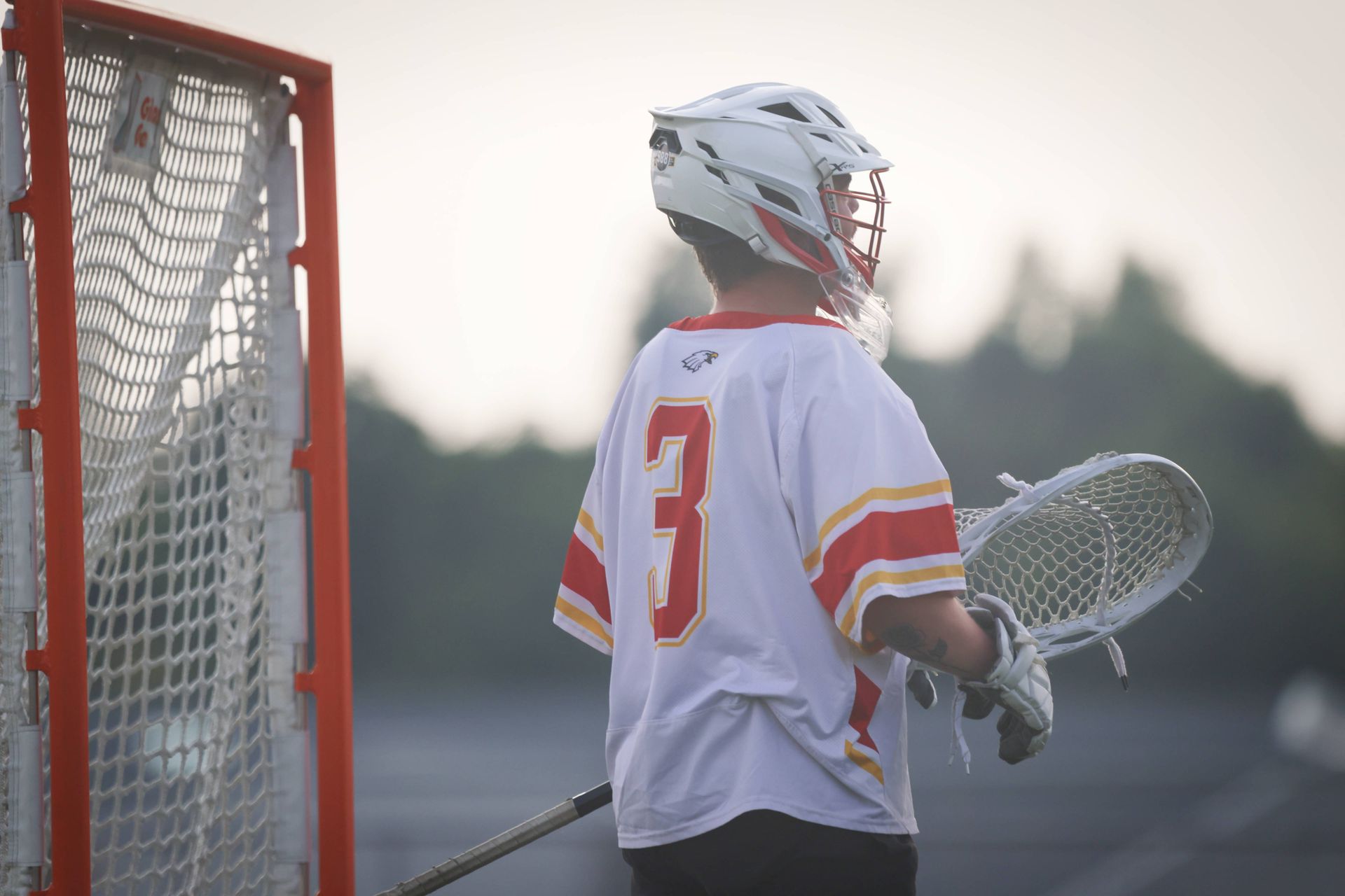 Lacrosse player in white jersey stands by goal at dusk, holding a stick on the field.