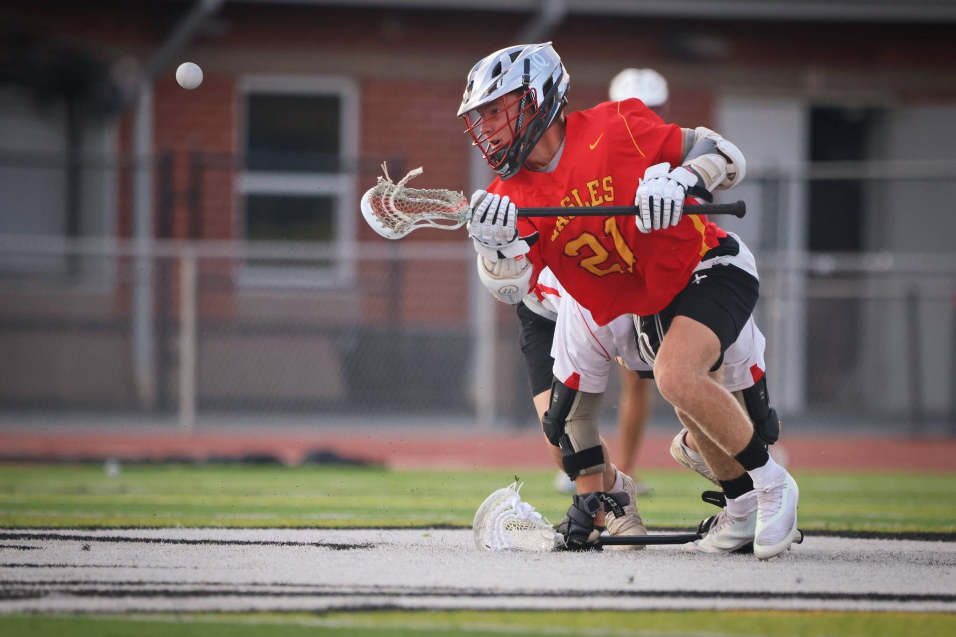 Two lacrosse players in action on a field, one in a red jersey holding the ball.