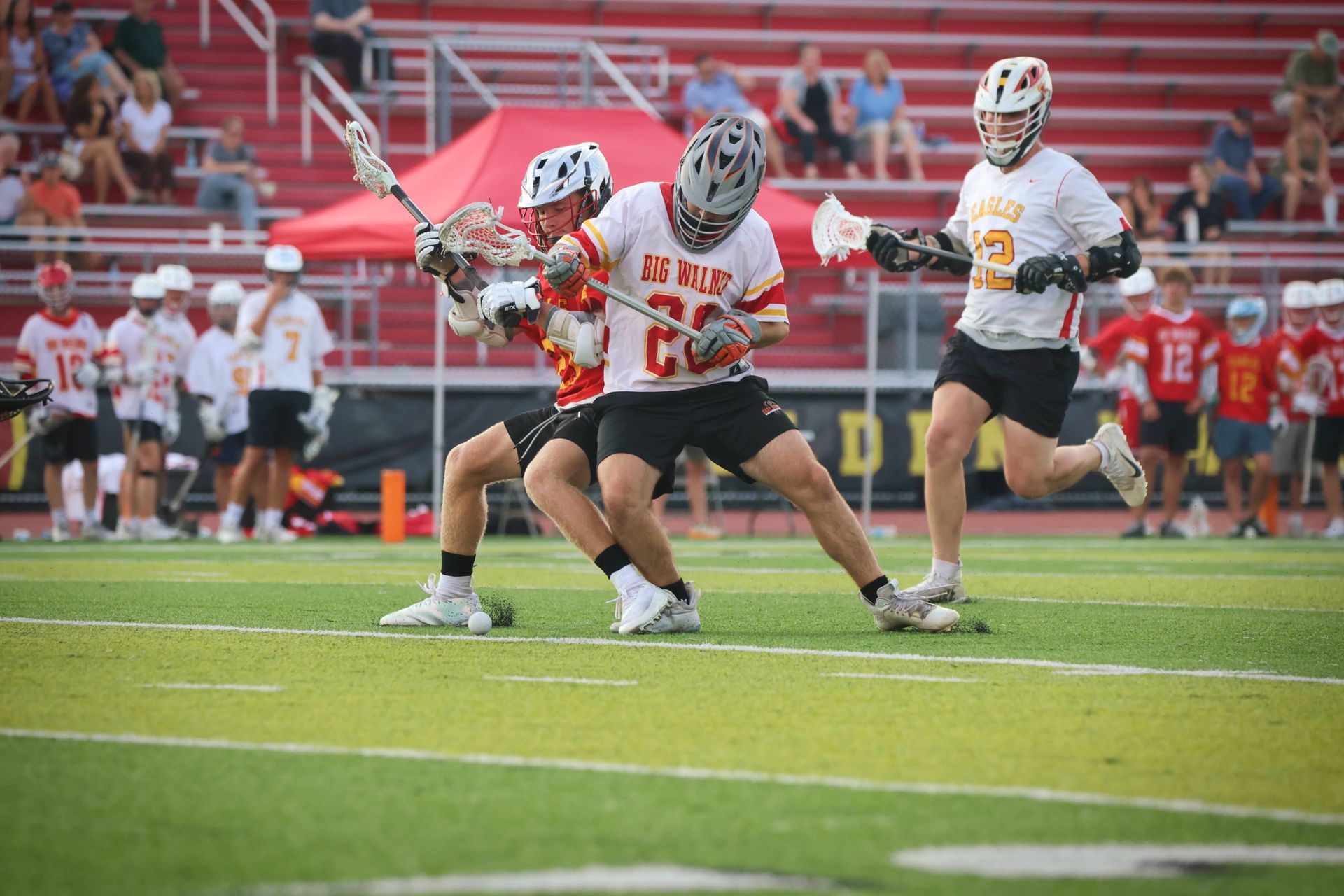Lacrosse players in white and red compete on a turf field with stadium seats in the background