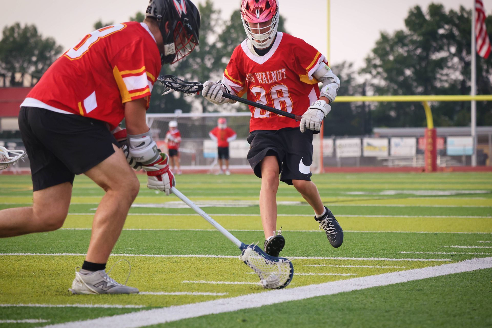 Two lacrosse players in red jerseys battle for the ball on a green turf field.