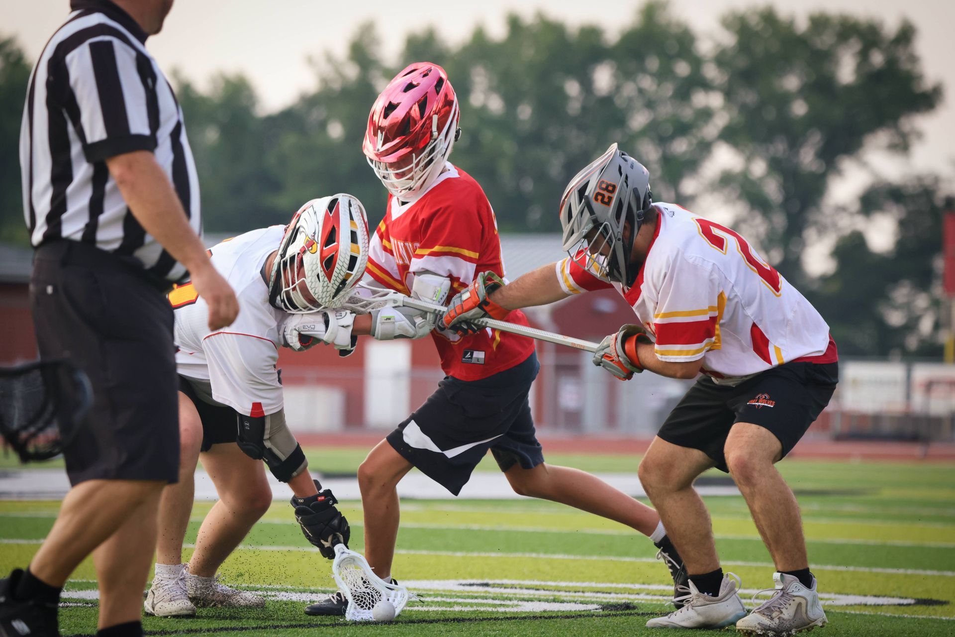 Three lacrosse players in red and white uniforms battle for the ball on a field