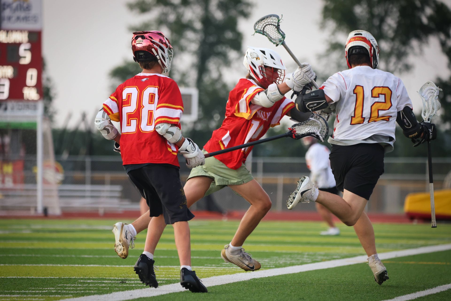Three lacrosse players in red and white uniforms battling for the ball on a wet field