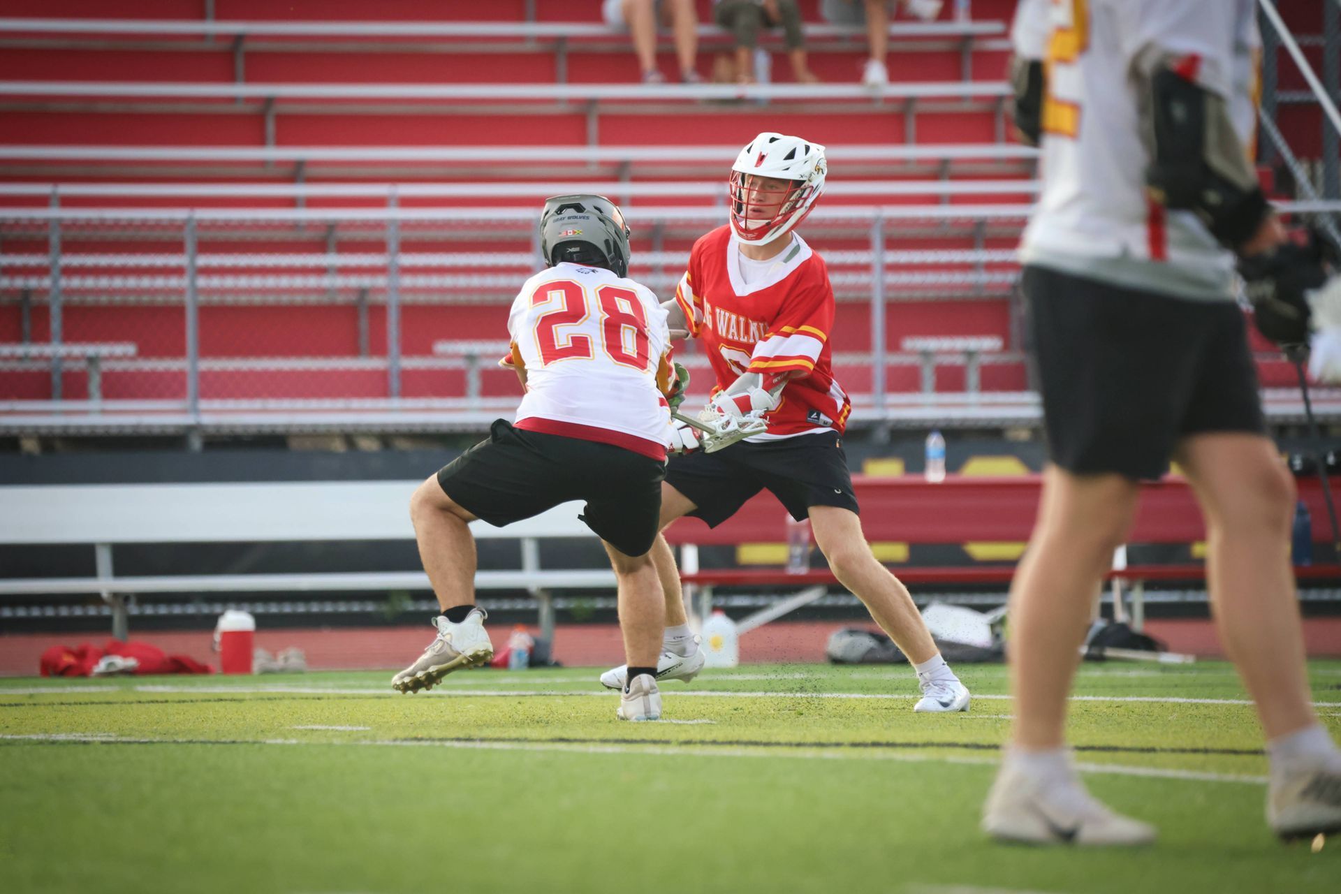 Two lacrosse players in red and white compete on a grassy field with stadium seats in the background.