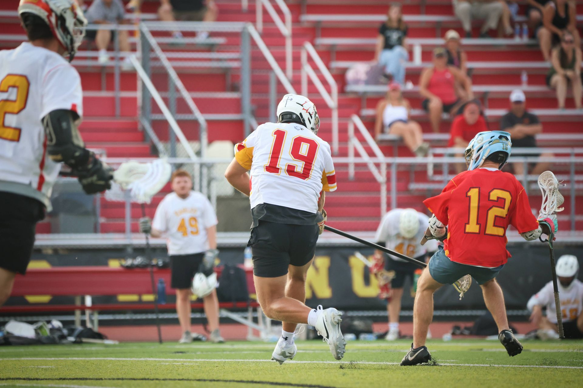 Lacrosse players in red and white uniforms sprint across a field during a game with bleachers in the background