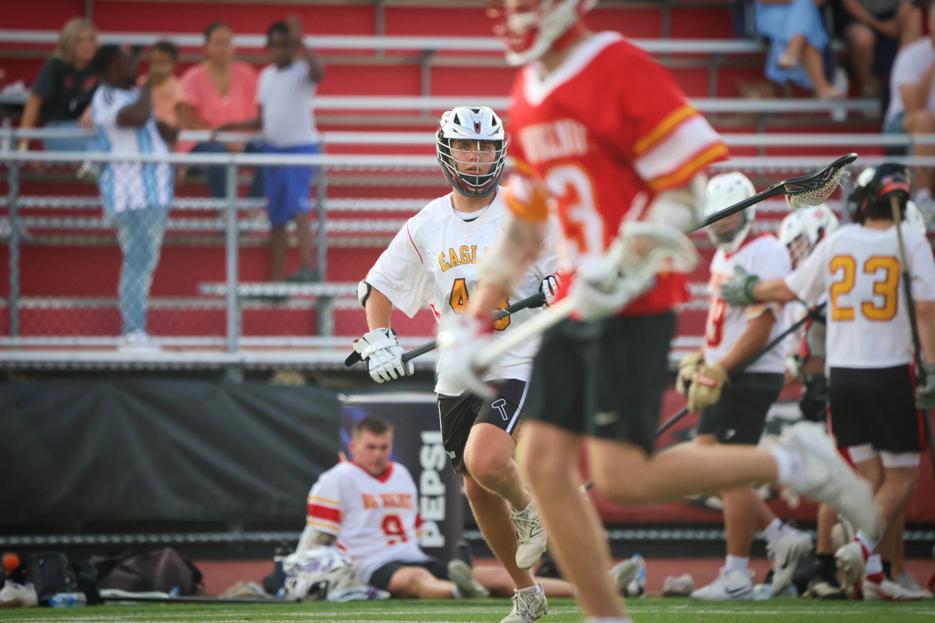 Lacrosse players in white and red uniforms on a field, with one player in focus and spectators in the stands.