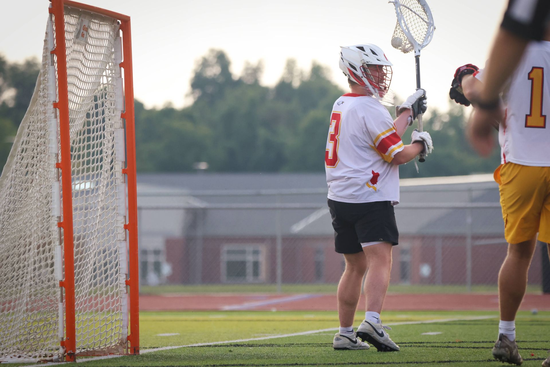 Lacrosse goalie in white uniform stands by the goal on a field at dusk.