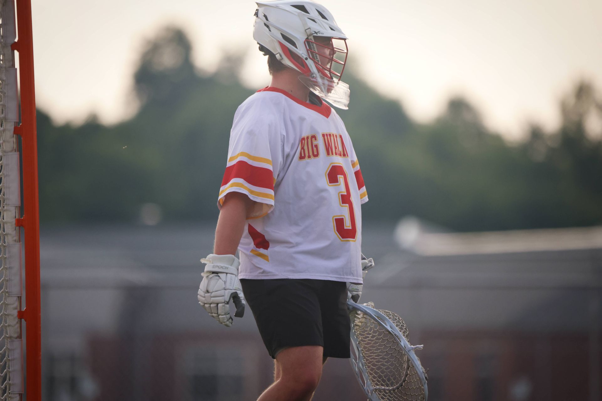 Lacrosse player in white and red uniform stands on the field holding a lacrosse stick at sunset