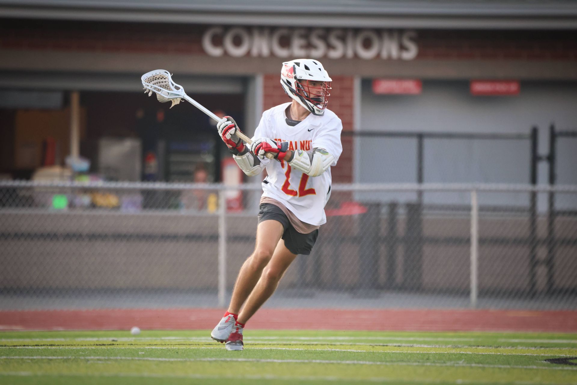 Lacrosse player in white uniform runs on a field with stick raised in front of a concession stand.