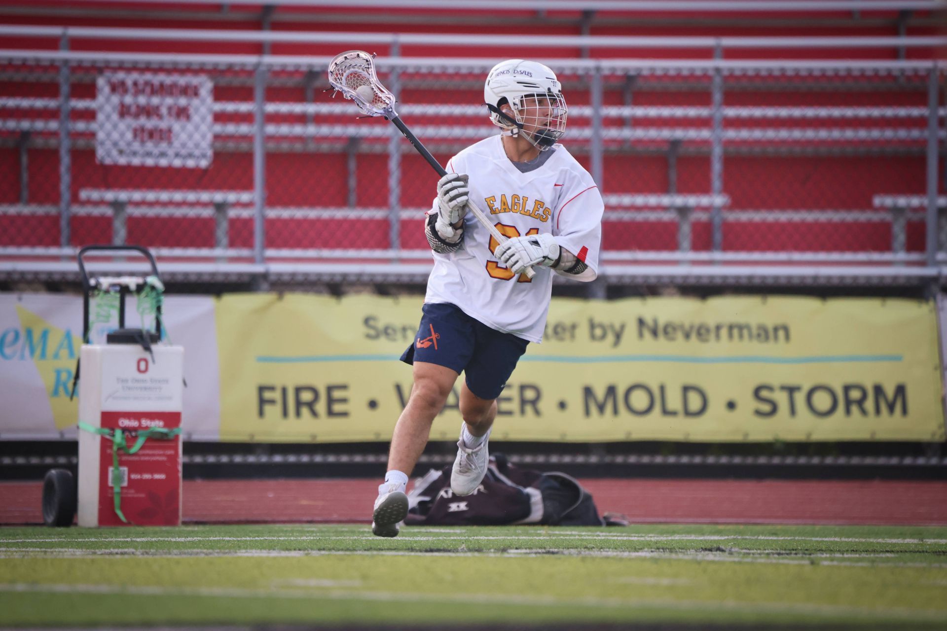 Lacrosse player in white jersey holds a stick on a field with red stadium bleachers behind him.