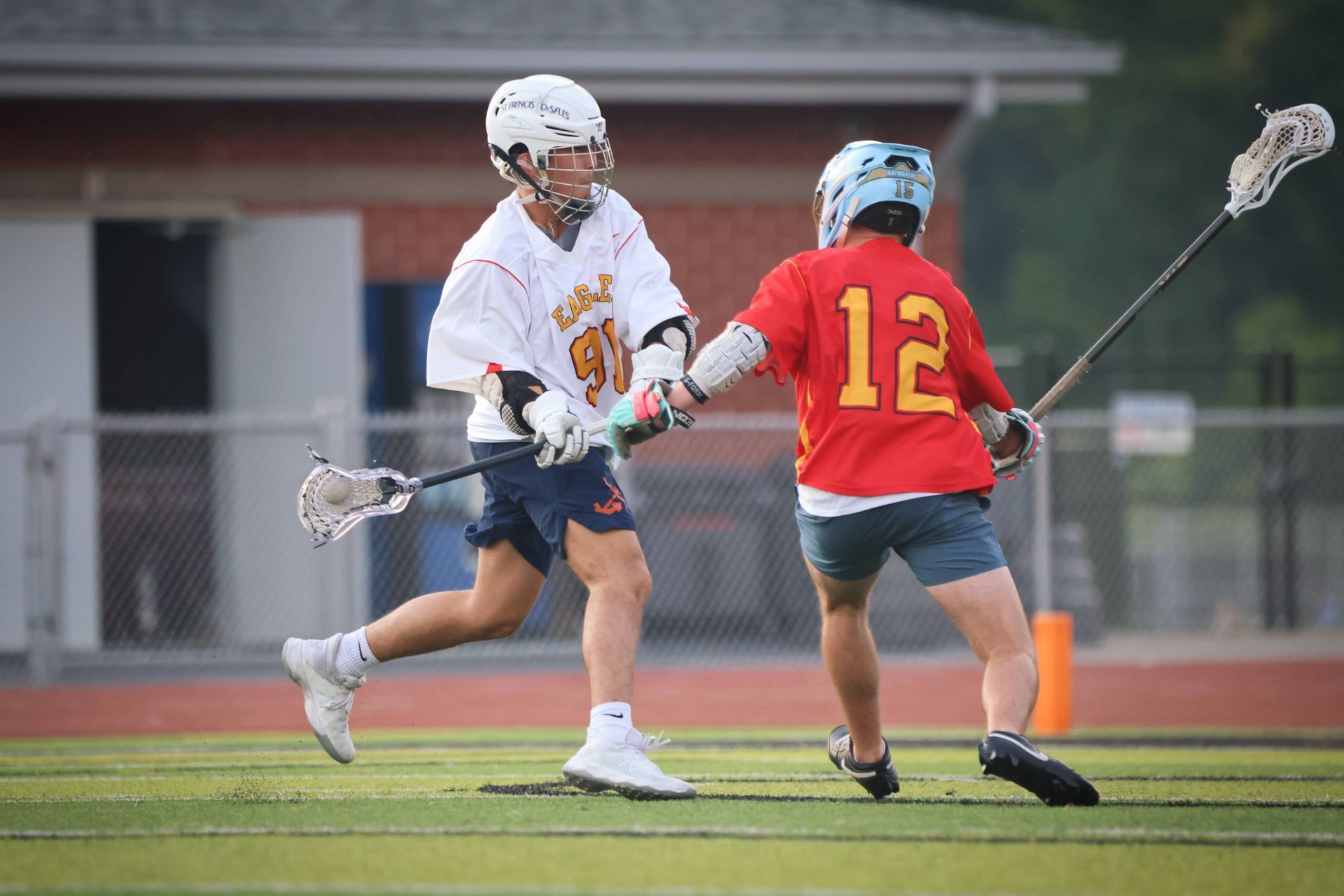 Two lacrosse players compete on a field, one in white and one in red with number 12.