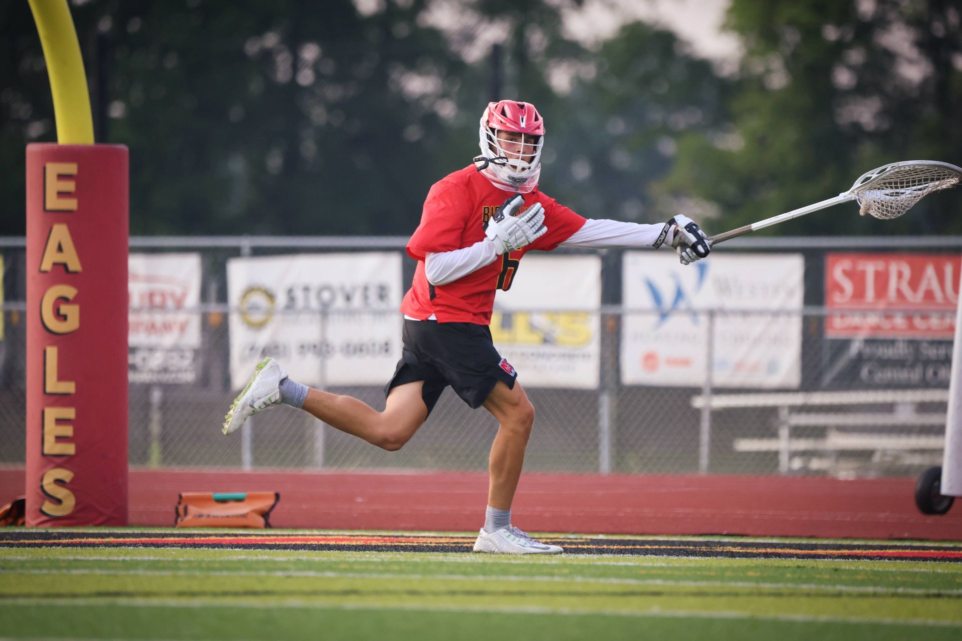 Lacrosse player in red and white sprints on a field, mid-shot, near an “EAGLES” goalpost banner.