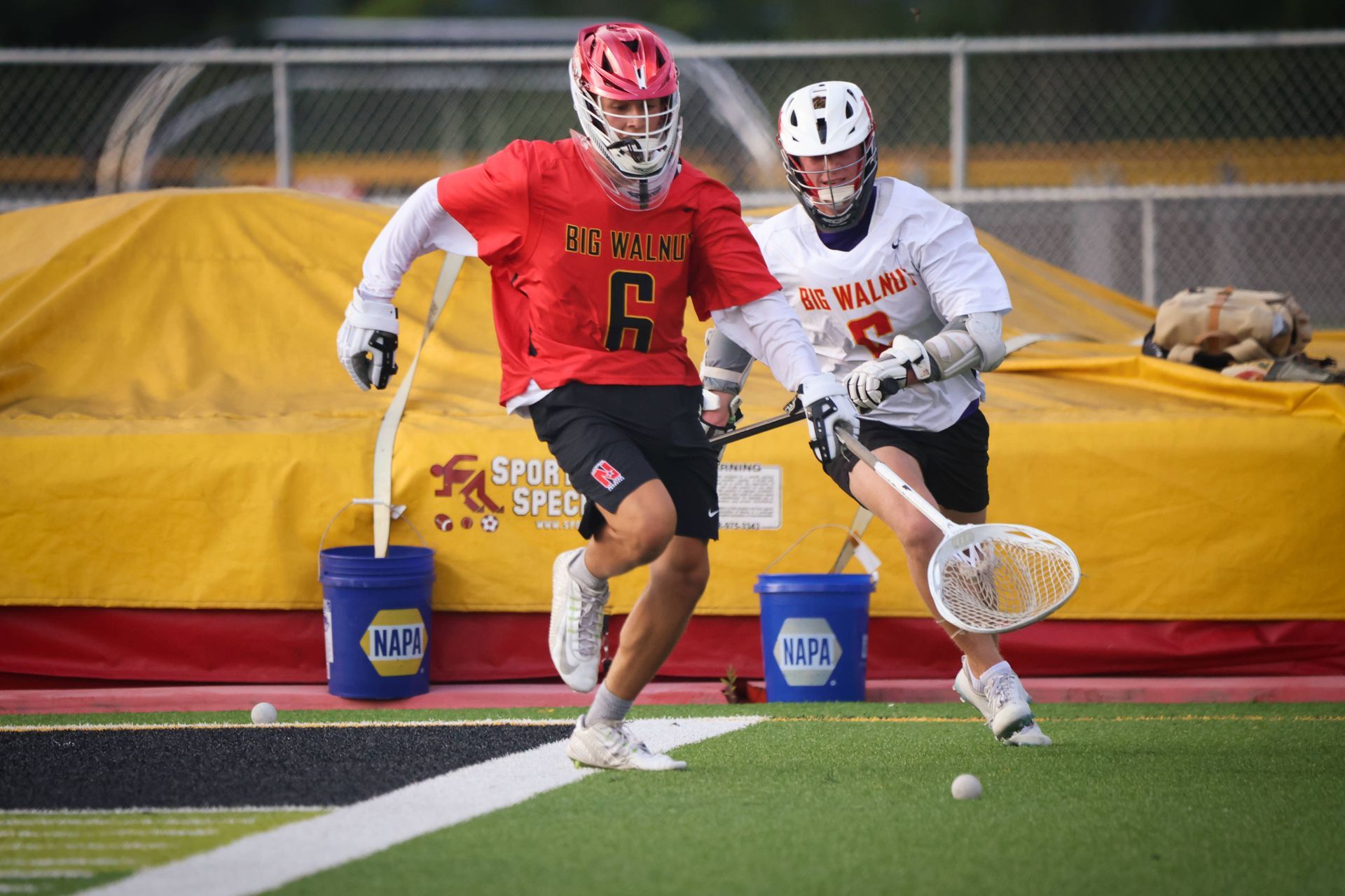 Two lacrosse players in red and white chase a loose ball on the field during a game.