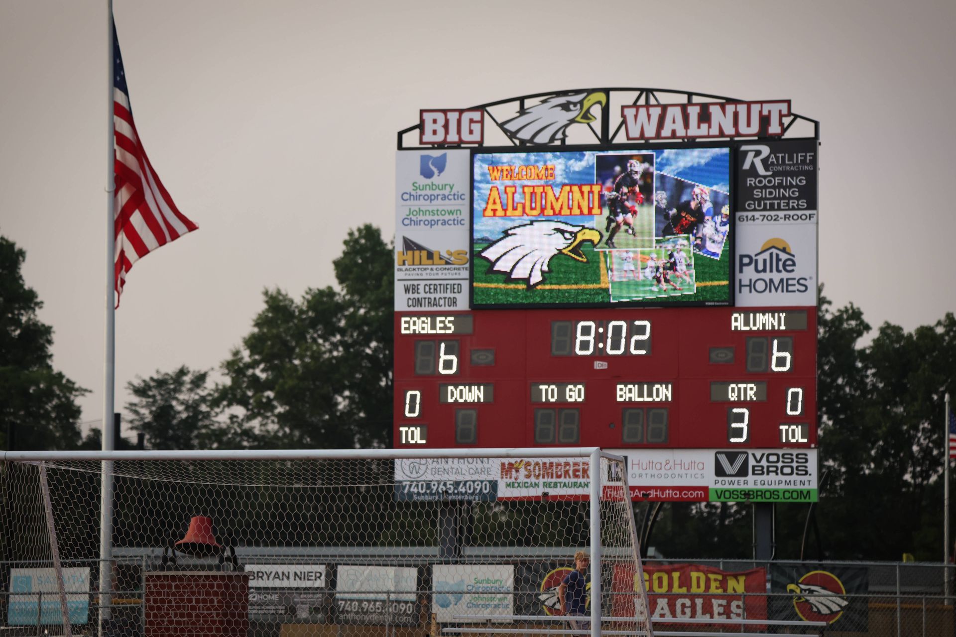 High school lacrosse scoreboard at dusk, showing 8:02 and a 6-6 score, with an American flag nearby.