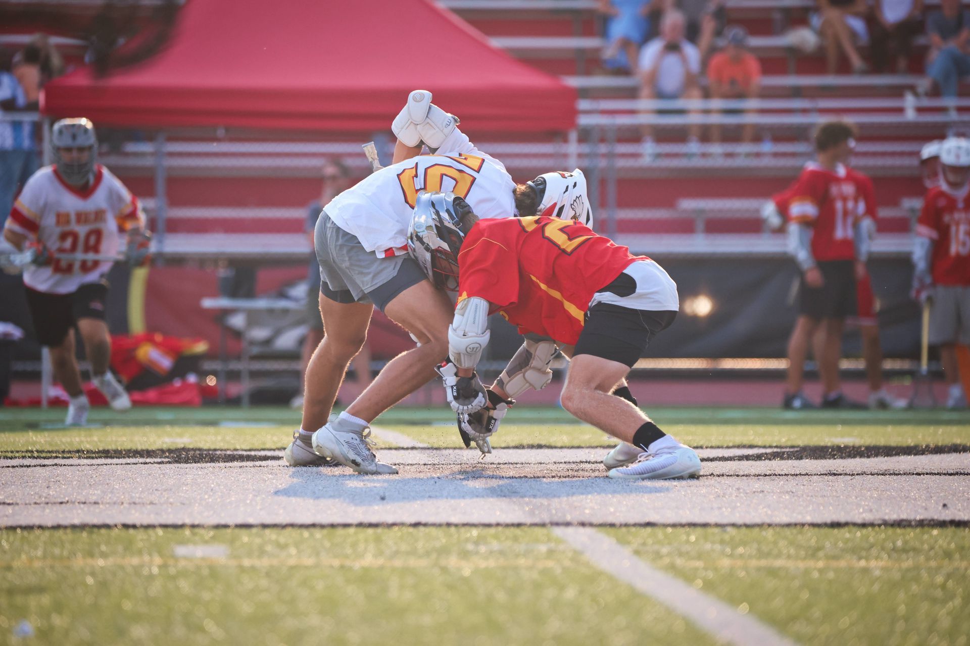 Two lacrosse players crouch for a faceoff on a sunlit field near the sideline.