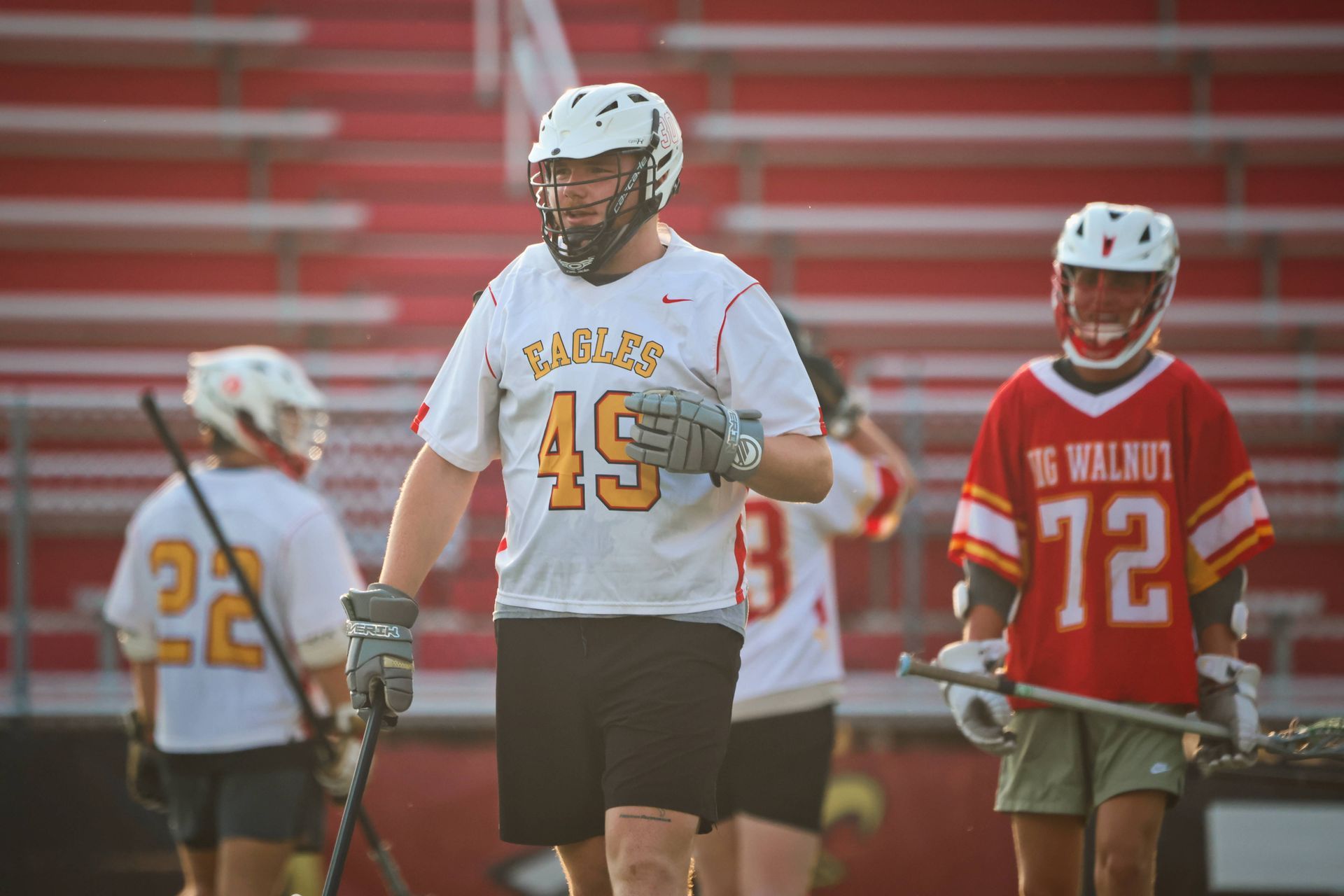 Lacrosse players in white and red uniforms on a field, with bleachers in the background.