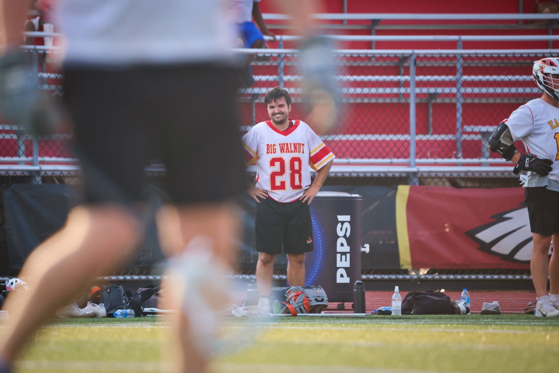 Lacrosse player in white and red jersey stands on field behind blurred foreground player