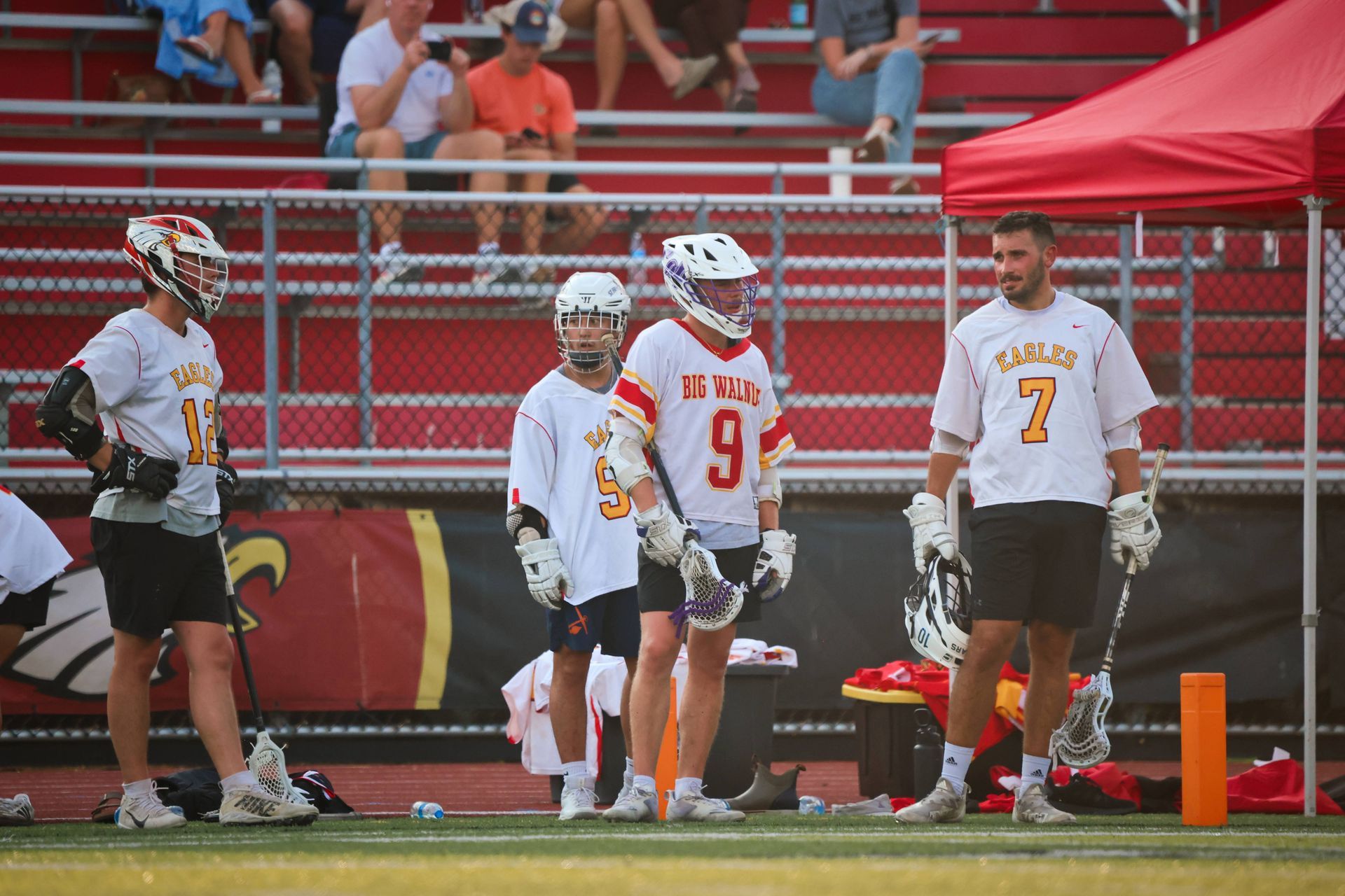 Lacrosse players in white and orange uniforms stand on the sideline near a red canopy on a field.