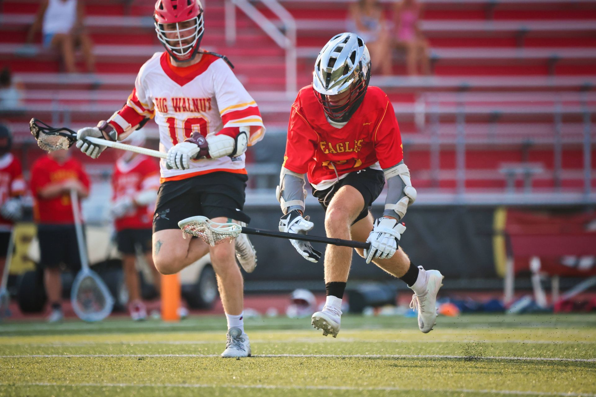 Two lacrosse players in red and white uniforms sprint on a field during a game.