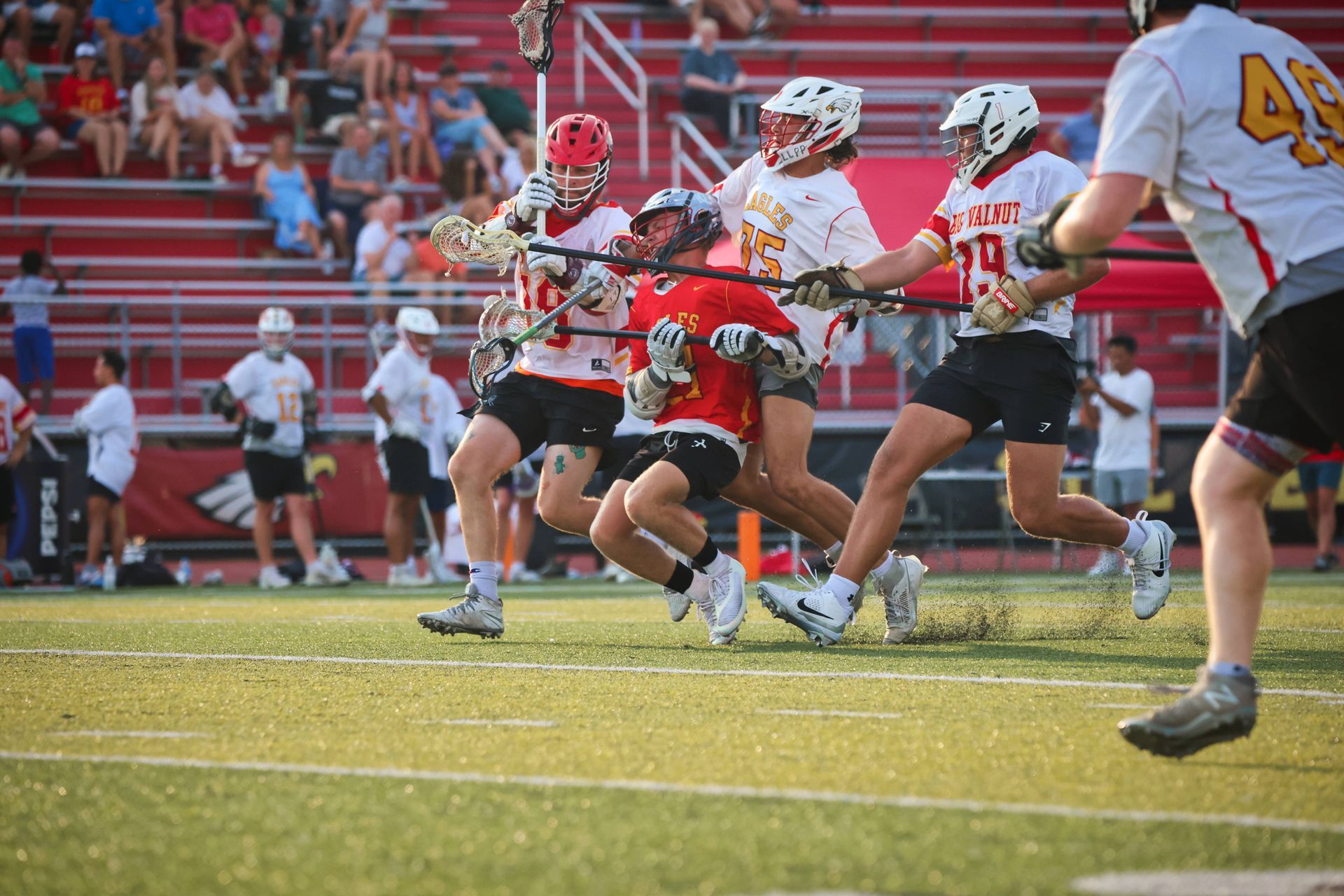 Lacrosse players in red and white sprint on a field during a game, with spectators in the stands.
