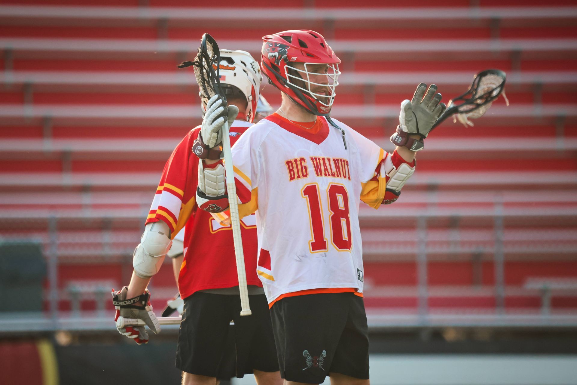 Two lacrosse players in red and white uniforms on a field, one holding a stick upright.