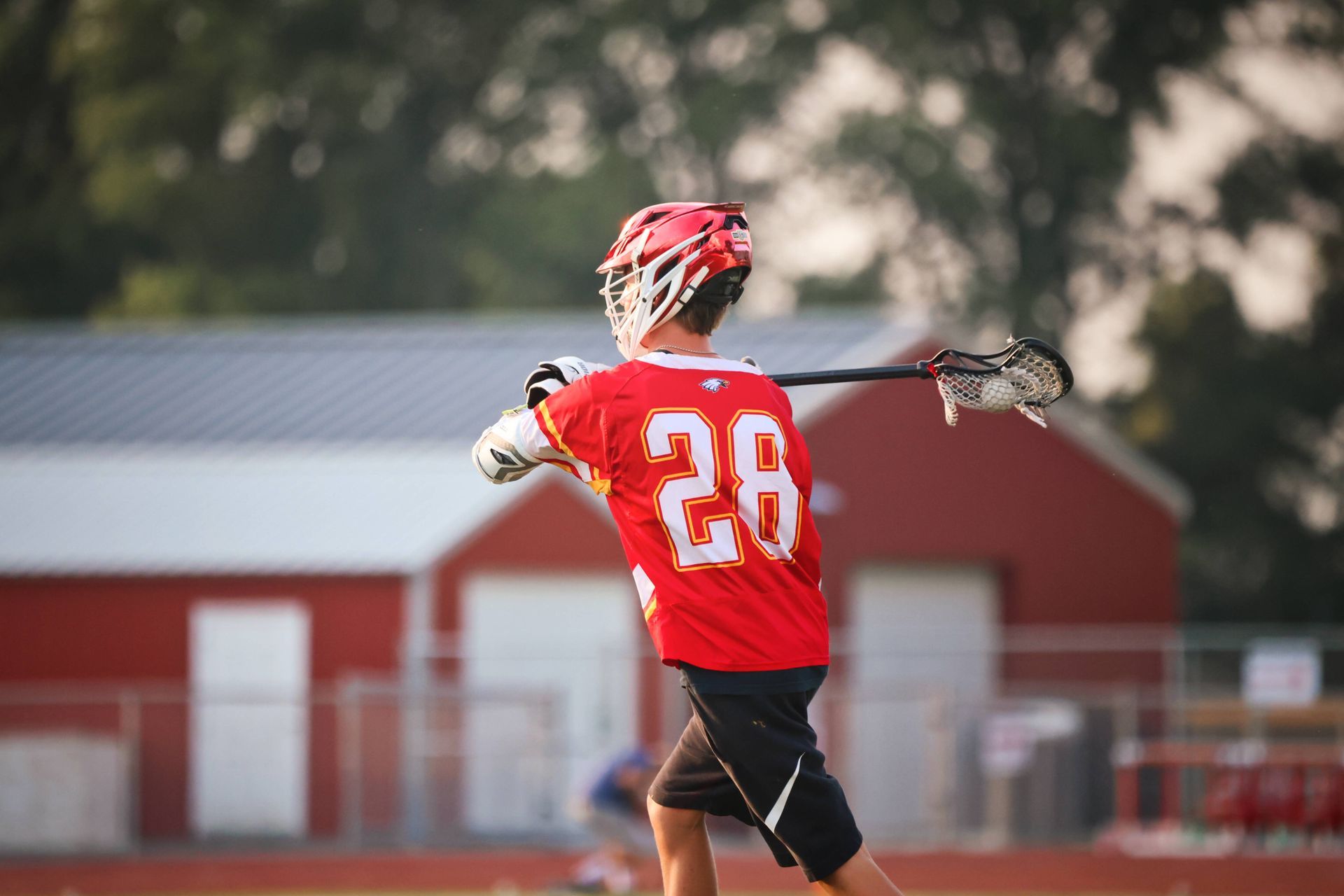 Lacrosse player in red jersey number 28 swings a stick on a field.