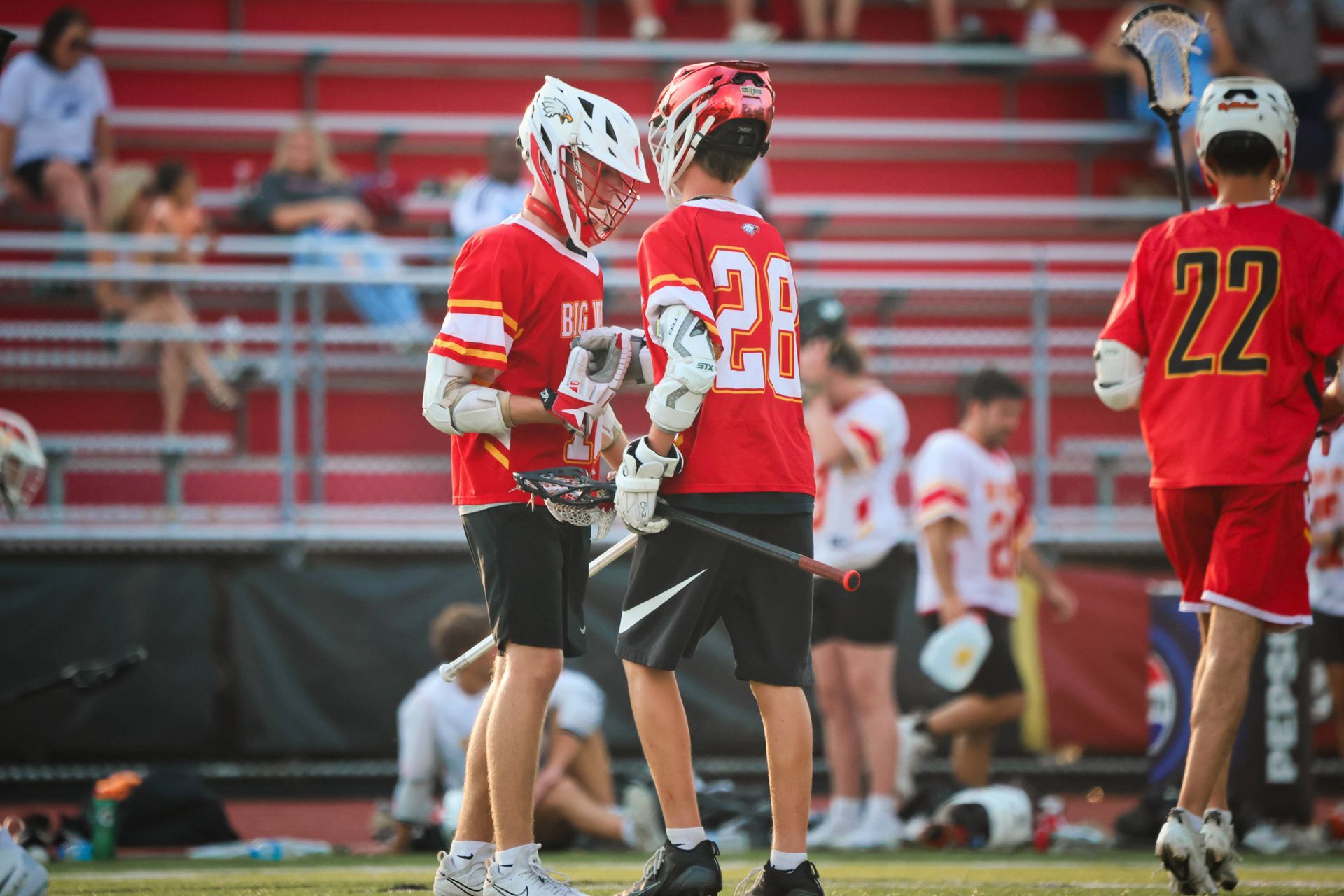 Lacrosse players in red uniforms stand on the field near the sideline during a game.