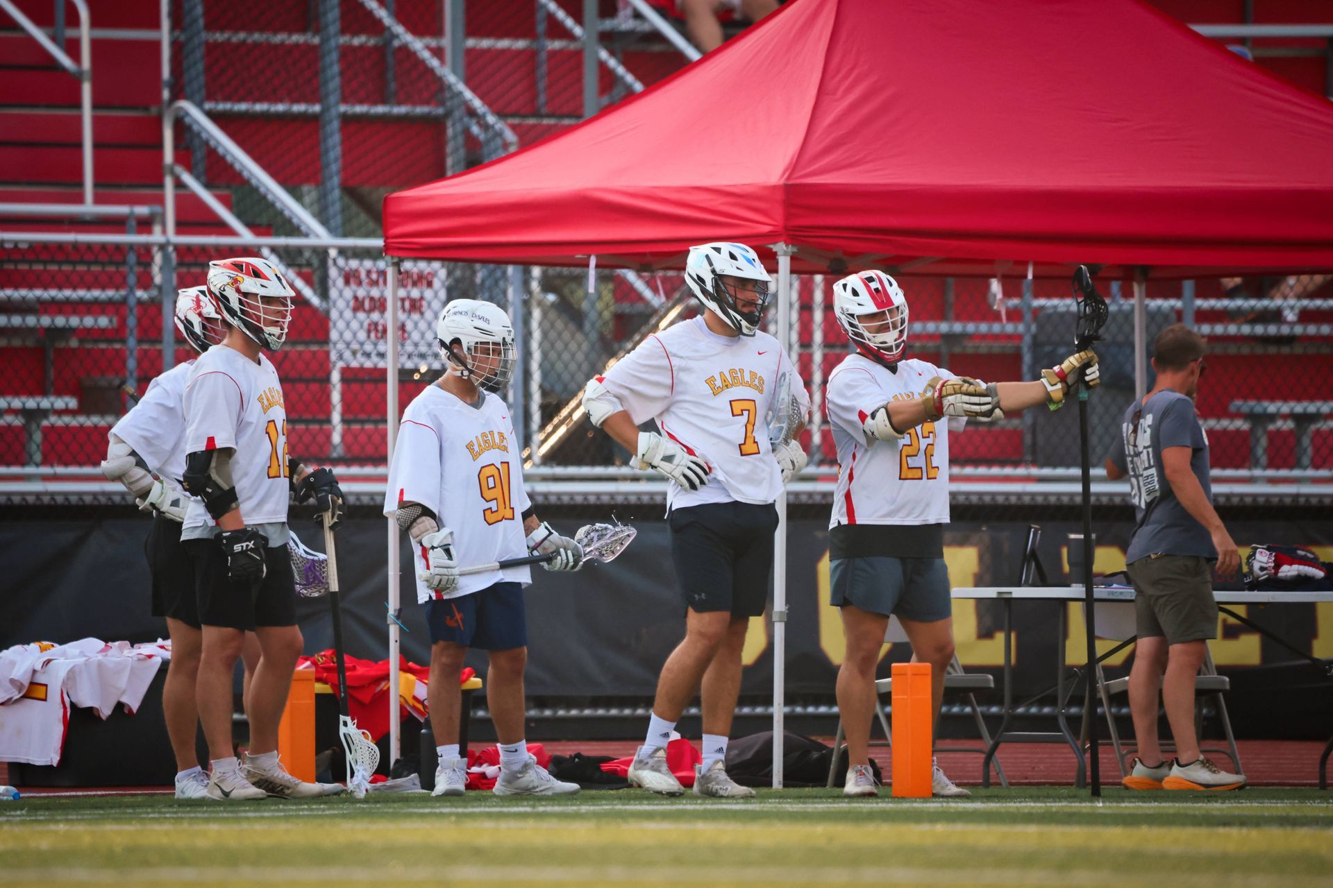 Lacrosse players in white jerseys stand by the sideline near orange cones on a field, with red bleachers behind them