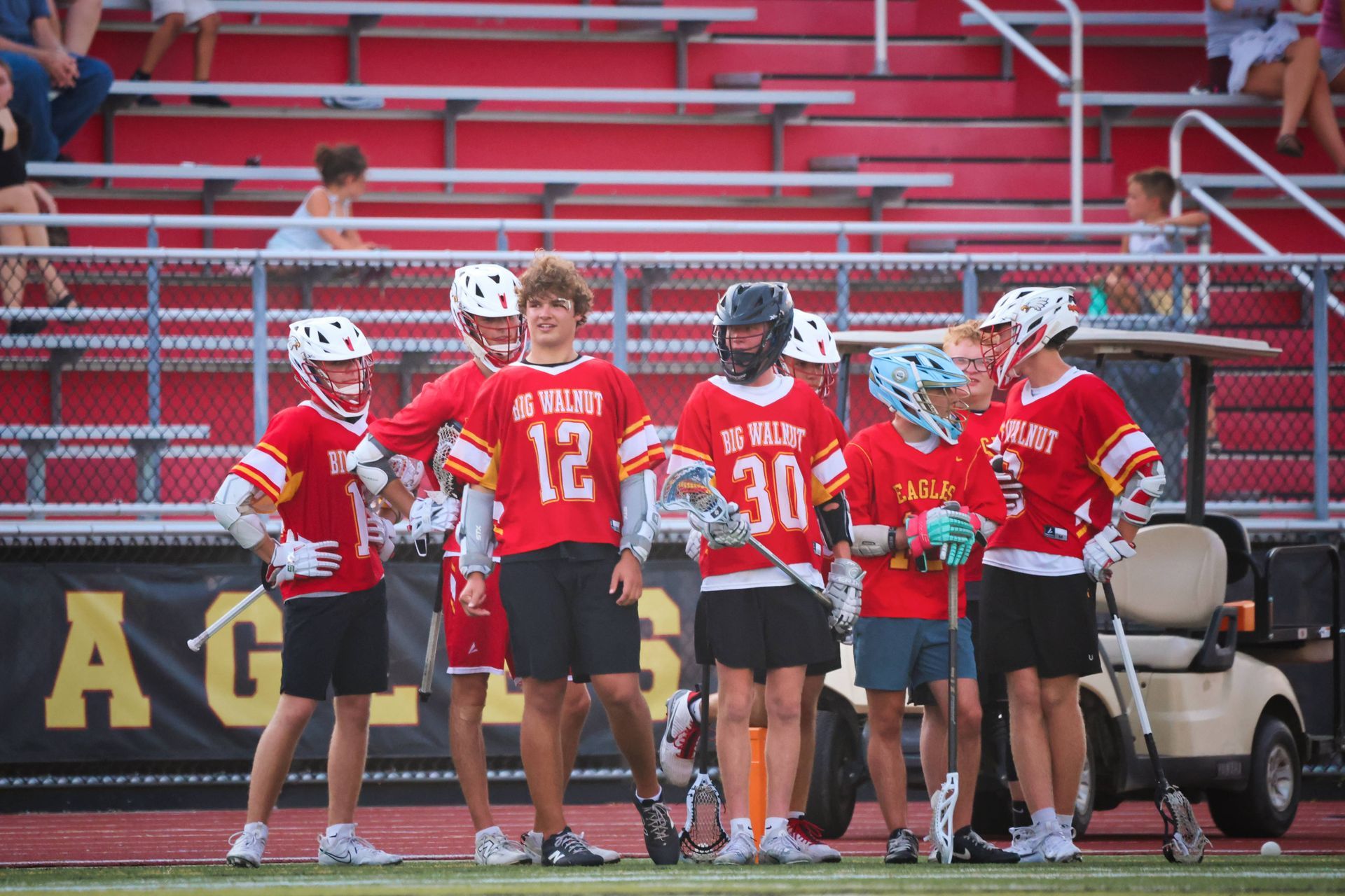 Lacrosse players in red jerseys huddle on the field near bleachers, with one coach standing beside them.